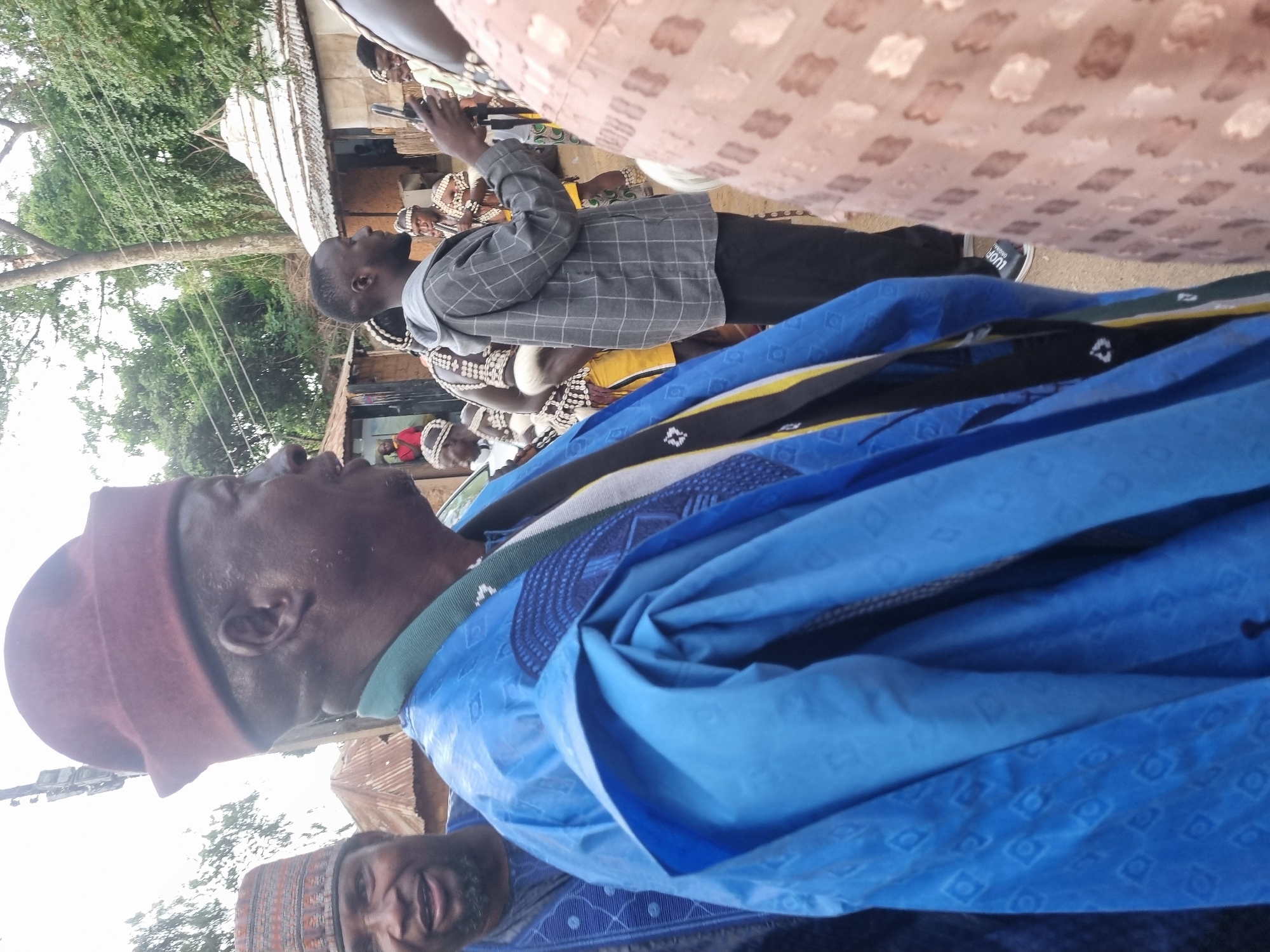 The image depicts a gathering of people outdoors, likely in a rural or semi-urban setting in Jos, Nigeria. In the foreground, a man wearing a traditional blue robe and a maroon cap is the central focus. He is looking to his right. To his left, another man in a patterned cap and blue attire is partially visible, also looking to his right. Behind them, a young man in a grey checkered jacket and black trousers is holding a mobile phone. Further in the background, several other individuals are present, some wearing traditional regalia with intricate beadwork and headpieces, and some appear to be playing musical instruments. Structures resembling huts or traditional buildings with thatched or corrugated iron roofs are visible in the background, along with trees and utility poles. The overall atmosphere suggests a community event or celebration.