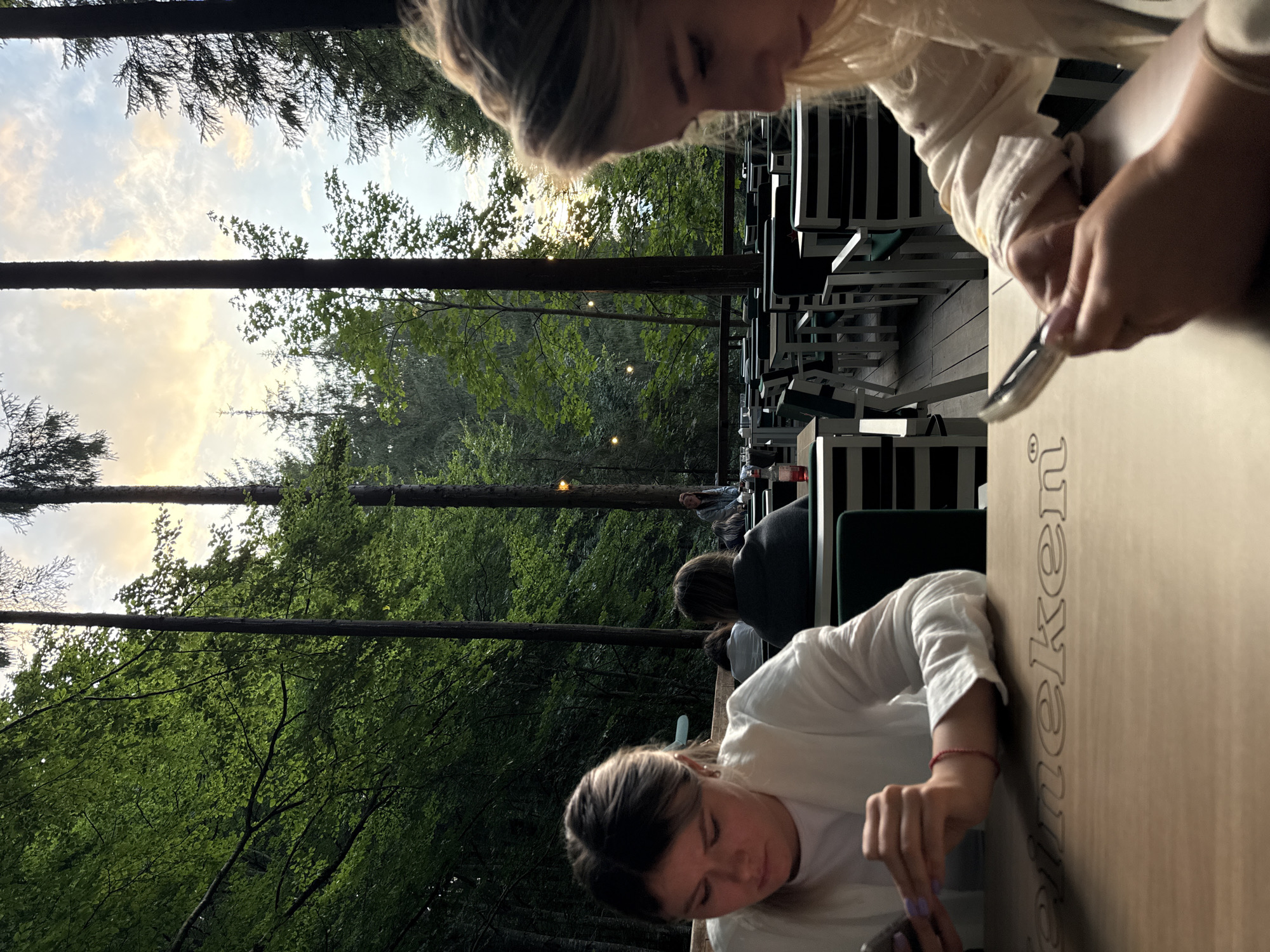 The image depicts an outdoor seating area in a forest setting, likely a restaurant or cafe, in Arefu, Romania. The foreground shows two women sitting at tables, both engaged with their mobile phones. The woman on the left, closer to the camera, is wearing a white top and is looking down at her phone. The woman on the right, slightly out of focus and in profile, is also looking at her phone, wearing a light-colored top. The tables have a wooden surface with carved lettering, partially visible, that appears to read "Heineken". In the background, other patrons are seated at tables and chairs, amidst tall trees and lush green foliage. The sky is visible through the trees, with clouds and hints of sunset light. String lights are also present, suggesting this is an evening scene. The overall atmosphere is relaxed and casual, with a focus on nature and outdoor dining.