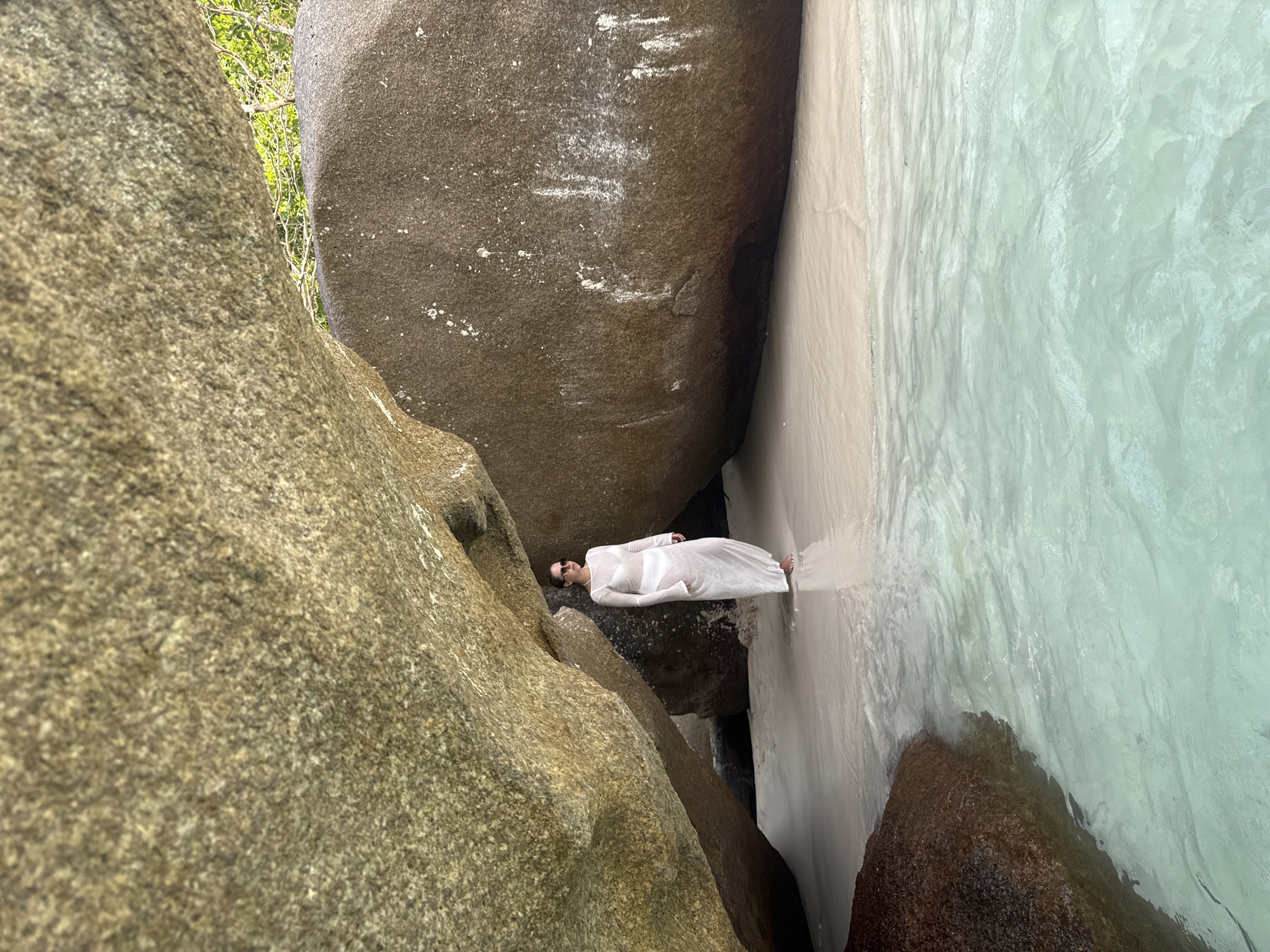 A vertical, medium-shot photograph captures a woman standing in shallow water between large rock formations. The woman, wearing a white dress and sunglasses, is positioned horizontally in the frame. To her left, a large, textured rock formation rises from the ground. To her right, another large rock formation meets the light-colored sand and turquoise water. The scene is set in the city of Lam Kaen, Thailand.