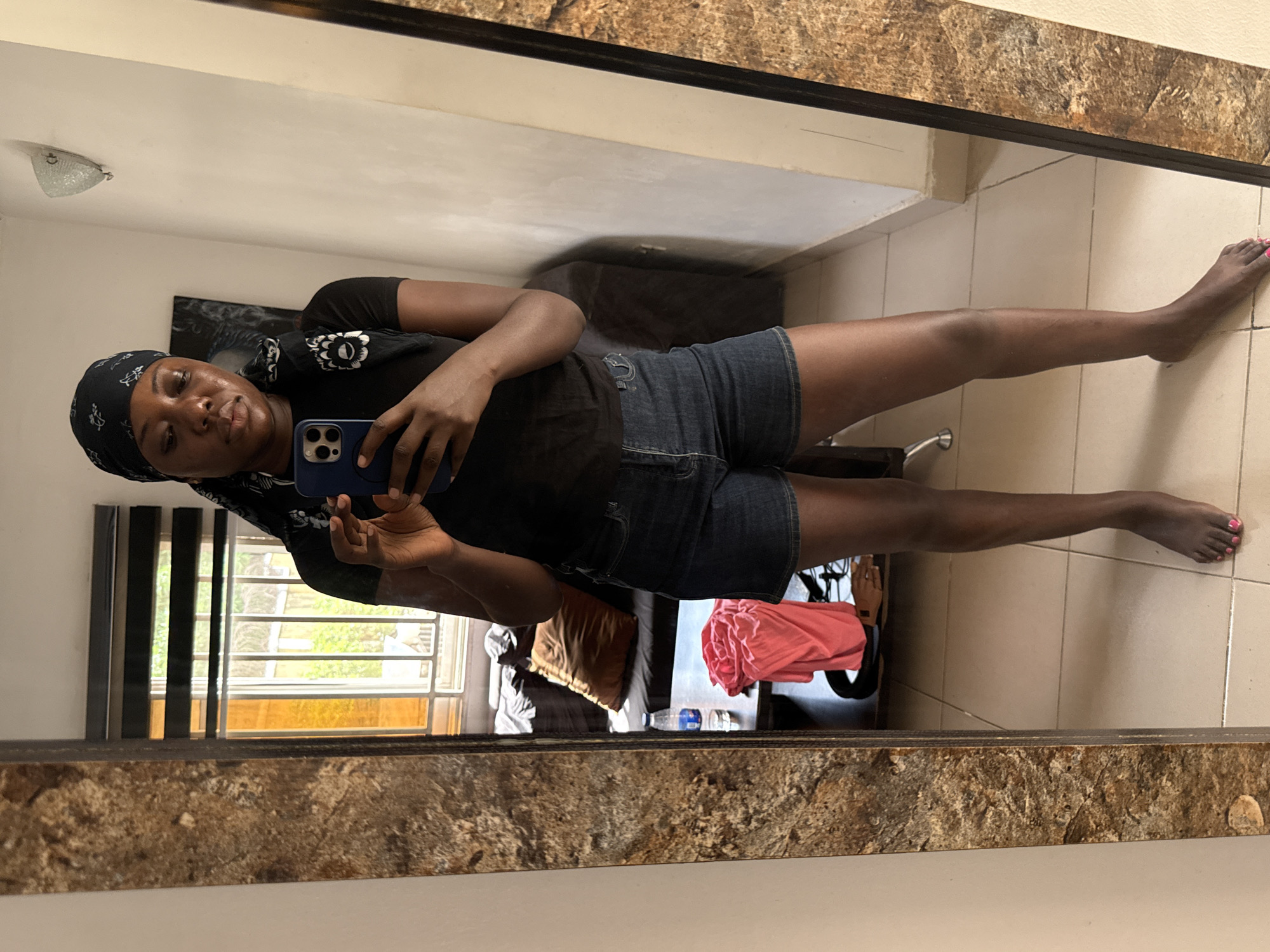 A dark-skinned woman with a black headscarf is taking a mirror selfie in what appears to be an apartment. She's wearing a black shirt and jean shorts. The apartment seems to be in Abuja, Nigeria, based on the general architectural style and interior decor, with neutral-toned walls and tile flooring. A window with partially closed blinds is visible, along with a bed with pillows, a water bottle and some clothing items. The mirror has a textured, marble-like frame.