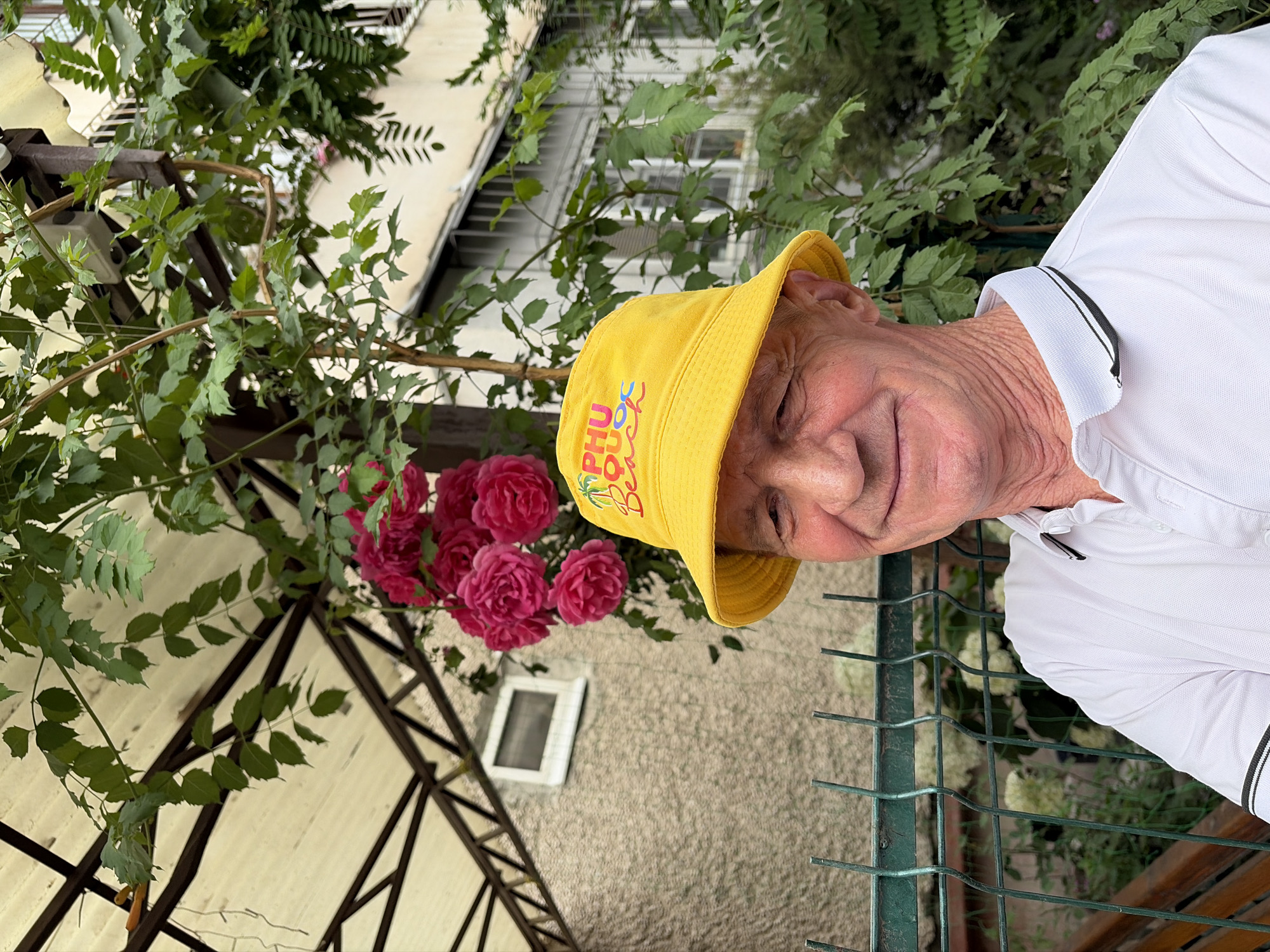 A man wearing a yellow bucket hat with the words "PHU QUOC BEACH" printed on it and a white polo shirt stands outdoors. He is smiling, and his face is wrinkled. Behind him, there is a bush with pink roses, a green fence, and a building. The man is looking at the camera.