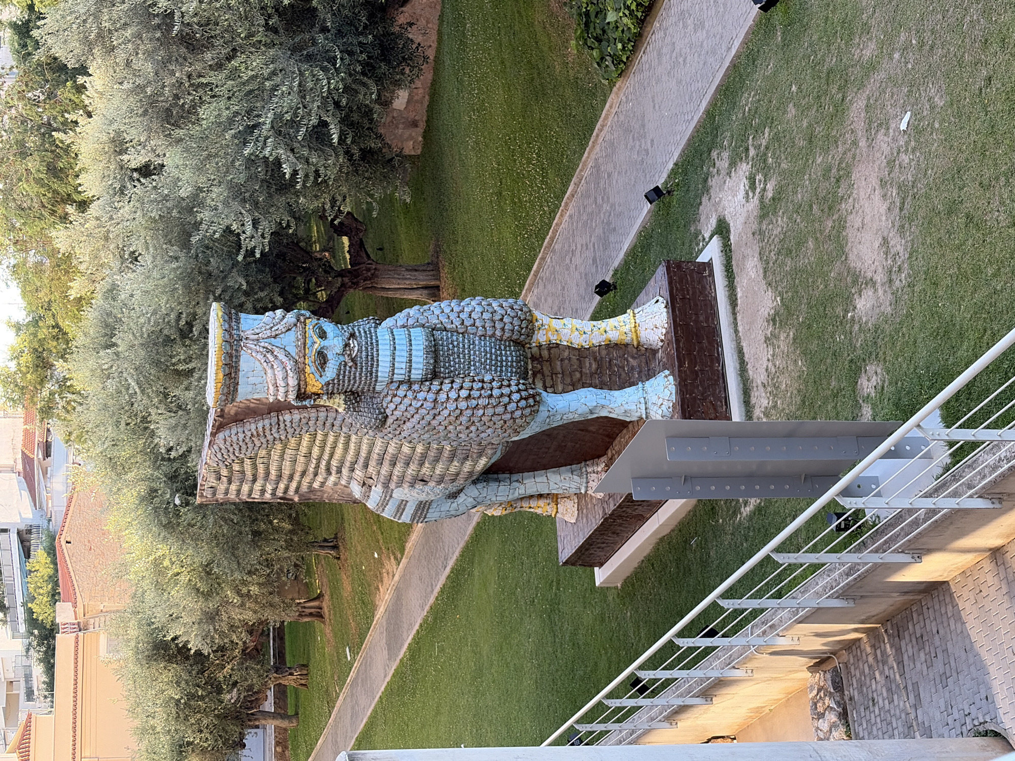 This media file captures a close-up, high-angle view of a large, mosaic-covered statue situated outdoors. The statue appears to be a stylized representation of a mythical creature or deity, possibly inspired by ancient Near Eastern art, given its scale and intricate design. It is predominantly blue and gold, with a patterned texture that suggests scales or feathers. The statue is mounted on a dark, rectangular base, which is itself elevated on a metal structure.
The setting is a well-maintained outdoor area, likely a park or garden. Lush green grass surrounds the statue, and a paved pathway runs alongside it. In the background, mature trees with full canopies are visible, suggesting a warm climate. To the left of the statue, a few residential buildings with tiled roofs can be seen, hinting at an urban or semi-urban environment. A modern metal railing and walkway are also prominent in the foreground, suggesting this image was taken from an elevated vantage point, possibly a balcony or walkway overlooking the statue.
The lighting suggests it is daytime, with a warm, soft glow, possibly late afternoon or early morning. The weather appears to be clear and pleasant. No people are visible in the frame. There is no discernible text in the image. The overall impression is one of public art in a serene, outdoor setting, with the statue as the central focus.