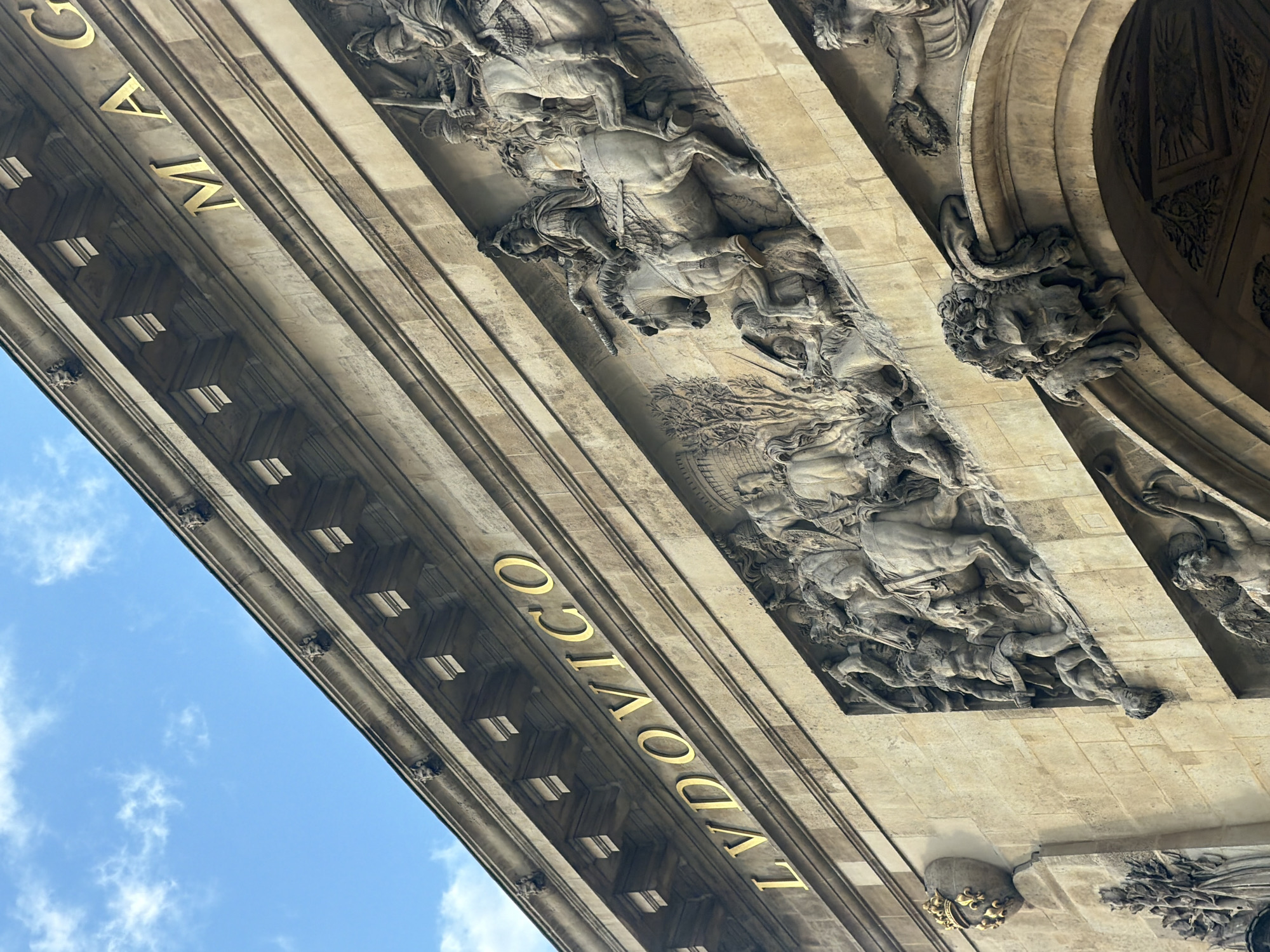 The photograph, taken from a low angle, captures a section of the Arc de Triomphe du Carrousel in Paris, France. The structure is primarily constructed of beige stone.
On the upper portions of the structure, high relief sculptures depict scenes of horses and riders. Below this, gilded lettering spells out "LUDOVICO". The bottom portion of the frame displays blue sky with wisps of white clouds.