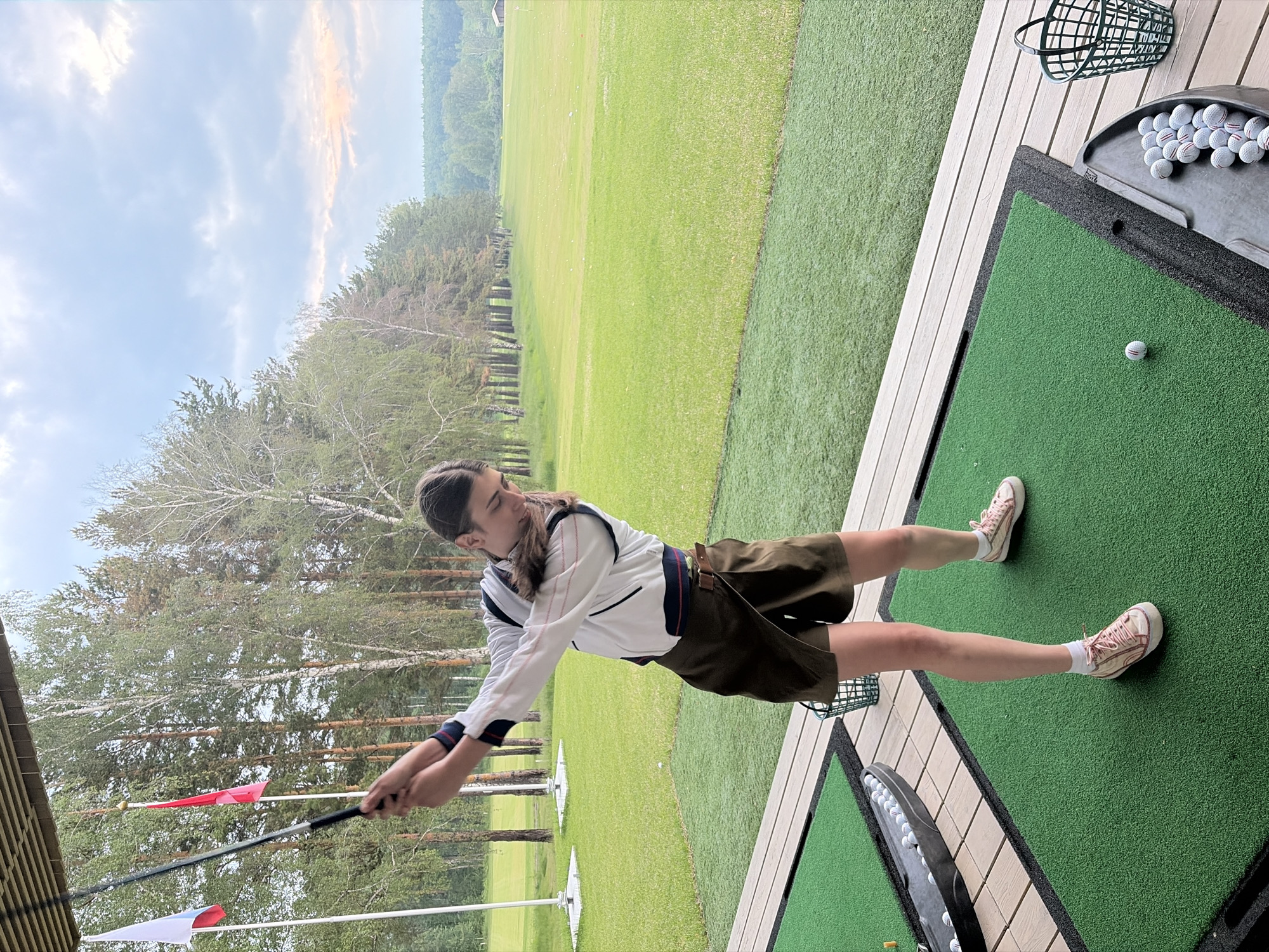 A young woman is captured in mid-swing on a golf driving range. She is positioned on a green mat, a golf club held in her hands, preparing to strike a golf ball. Her attire includes a white jacket with dark trim, a dark brown skort, and light pink sneakers. Behind her, a lush green field stretches towards a line of trees, with a partly cloudy sky overhead. Flags, one white and one red, are visible on poles to her left, suggesting the location is a sports facility. The environment includes a wooden deck and baskets of golf balls, indicating an outdoor recreational setting. The scene is observed from a slightly elevated perspective, looking down at the golfer and the range.