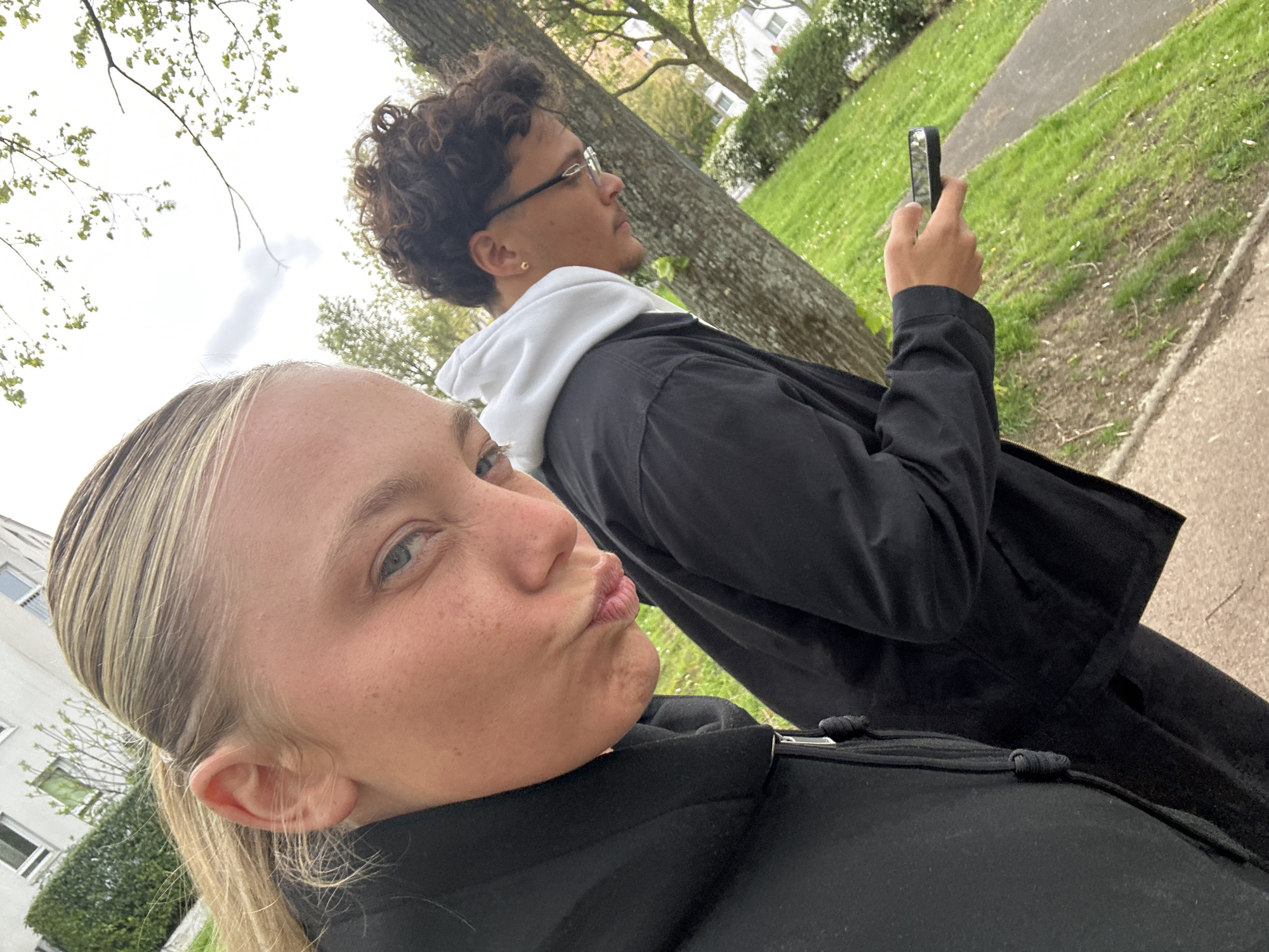 Two individuals are visible in the image. In the foreground, a fair-skinned woman with blonde hair pulled back in a ponytail is making a kissing face at the camera. Behind her is a male individual with curly hair, wearing glasses, a white hoodie, and a dark jacket, holding a cell phone. They appear to be outdoors in a park-like setting with green grass and trees. A building is visible in the background on the left side. The presence of trees lining the sidewalk is consistent with a urban landscape. Given the location specified (Noisiel, France), the buildings and foliage match known characteristics. They could be taking a self-portrait or recording something with the phone.