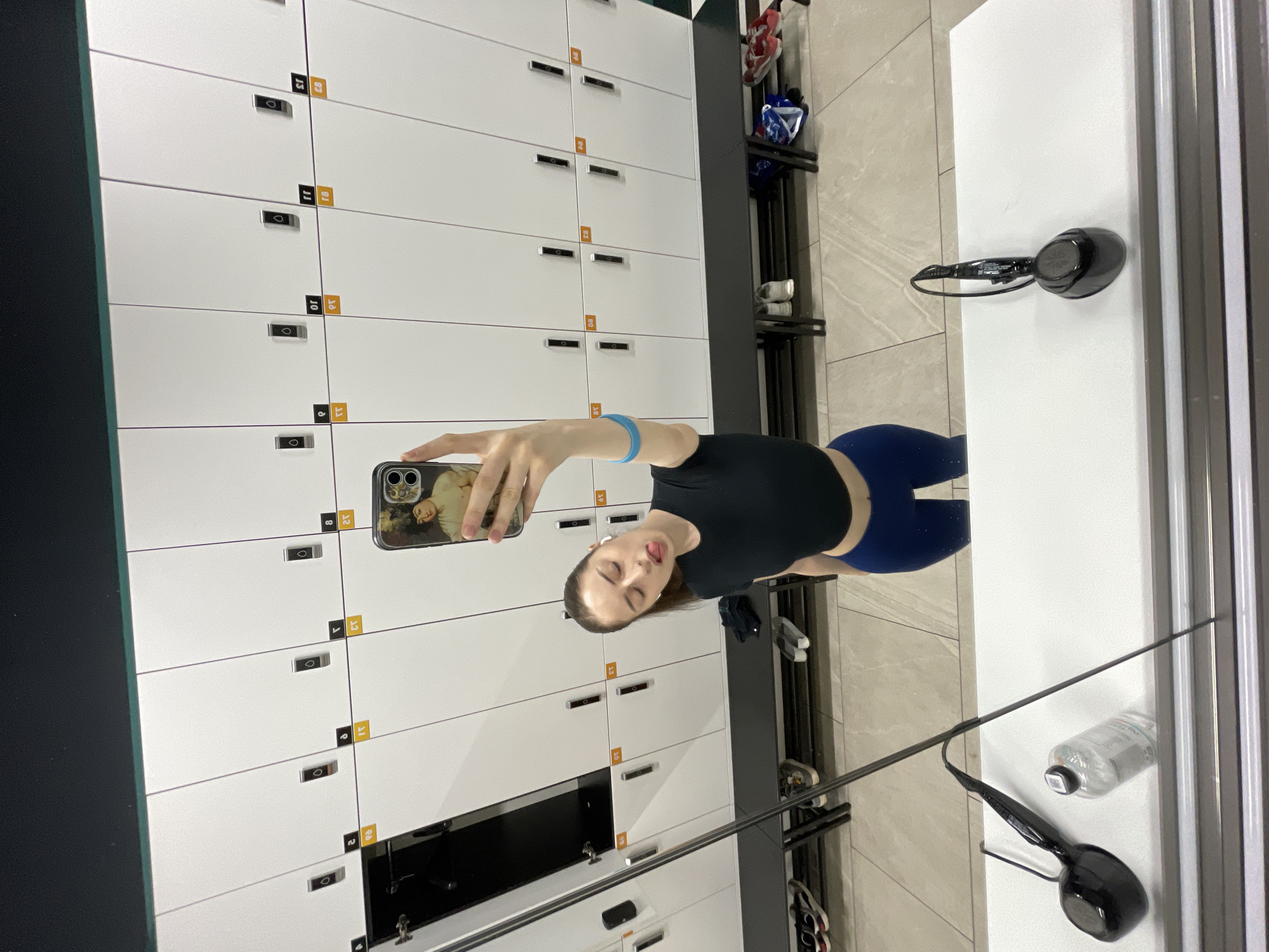 A young woman is taking a selfie in a gym locker room, with rows of white lockers in the background. She is wearing a black crop top and blue leggings. She has her tongue sticking out and is holding her phone with her right hand, while her left arm with a blue bracelet is bent at the elbow. A mirror reflects the woman and the locker room. In the foreground, on a white counter, are a pair of black headphones, a clear water bottle, and a thin metal pole. Below the lockers, on a black bench, are various shoes and bags. The floor is tiled with light grey and beige squares. The scene suggests a typical gym environment. The presence of lockers and workout attire implies a place for physical activity. The overall atmosphere is casual and personal, as indicated by the selfie and the woman's pose.