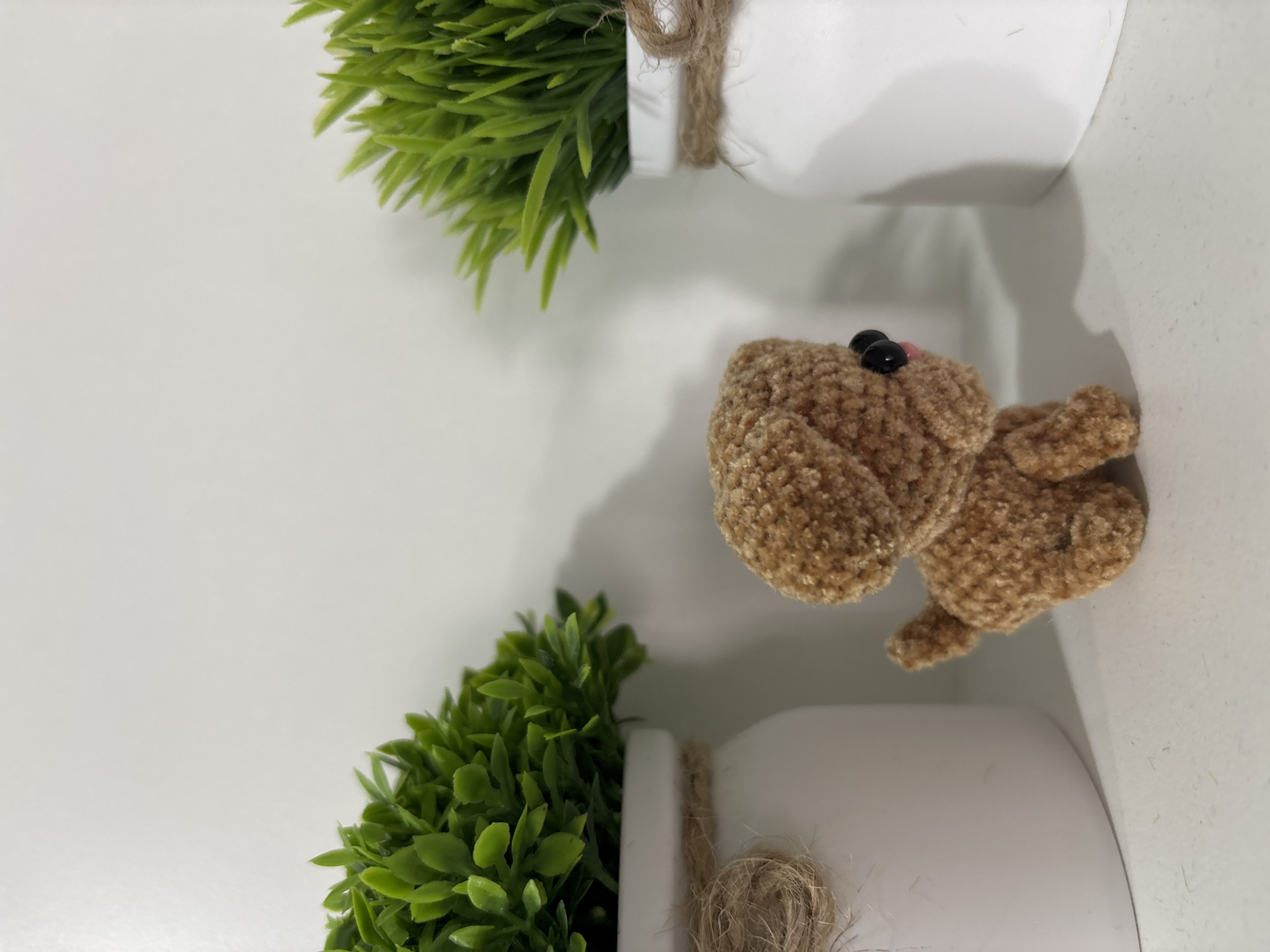 The scene, located in Izhevsk, Russia, depicts a small, light brown, crocheted dog plush toy positioned centrally on a white horizontal surface. The toy faces right, exhibiting black safety eyes and a small pink nose or mouth detail. Two white cylindrical pots containing artificial green foliage flank the plush toy. The pot on the left is partially visible and has a jute twine wrap around its rim, forming a knot on the left side. The pot on the right is fully visible, also with a jute twine wrap around its rim. The background is a plain white vertical surface. Shadows are cast behind the objects onto both the horizontal and vertical surfaces. No people are present, and no actions or interactions are depicted.