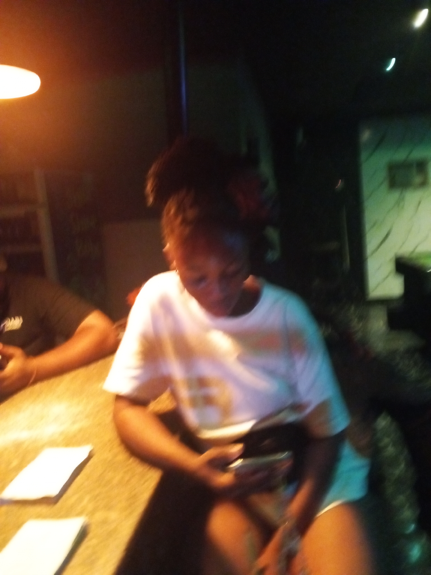 The image appears to be taken indoors, possibly in a bar or lounge due to the low lighting and the presence of a bar counter. A woman is sitting at the bar, looking down at a mobile phone she is holding in her hands. She is wearing a white t-shirt and appears to be wearing shorts. A man is partially visible to her left, also sitting at the bar counter. The counter has a paper or card on it. The location is unknown.