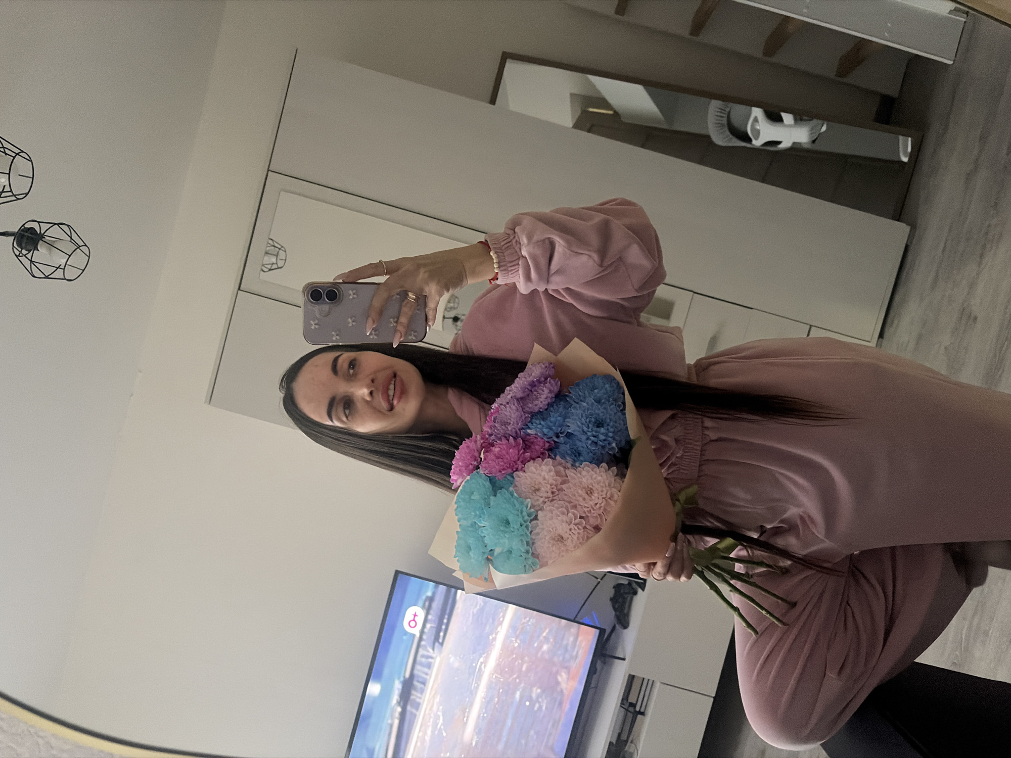 A fair-skinned woman is posing for a selfie in front of a mirror, holding a bouquet of multi-colored flowers. She has long, dark hair, and is wearing a pink two-piece lounge set. Her phone has a light purple case with white floral designs. The bouquet consists of pink, purple, and blue flowers wrapped in tan paper. Behind her is a television displaying a scene with water, possibly a beach or coastal area. The room has white walls and minimalist decor. A second mirror is visible in the background, reflecting a cleaning apparatus on a shelf. It is not possible to determine the location in the city of Saint Petersburg, Russia, based on the given image.