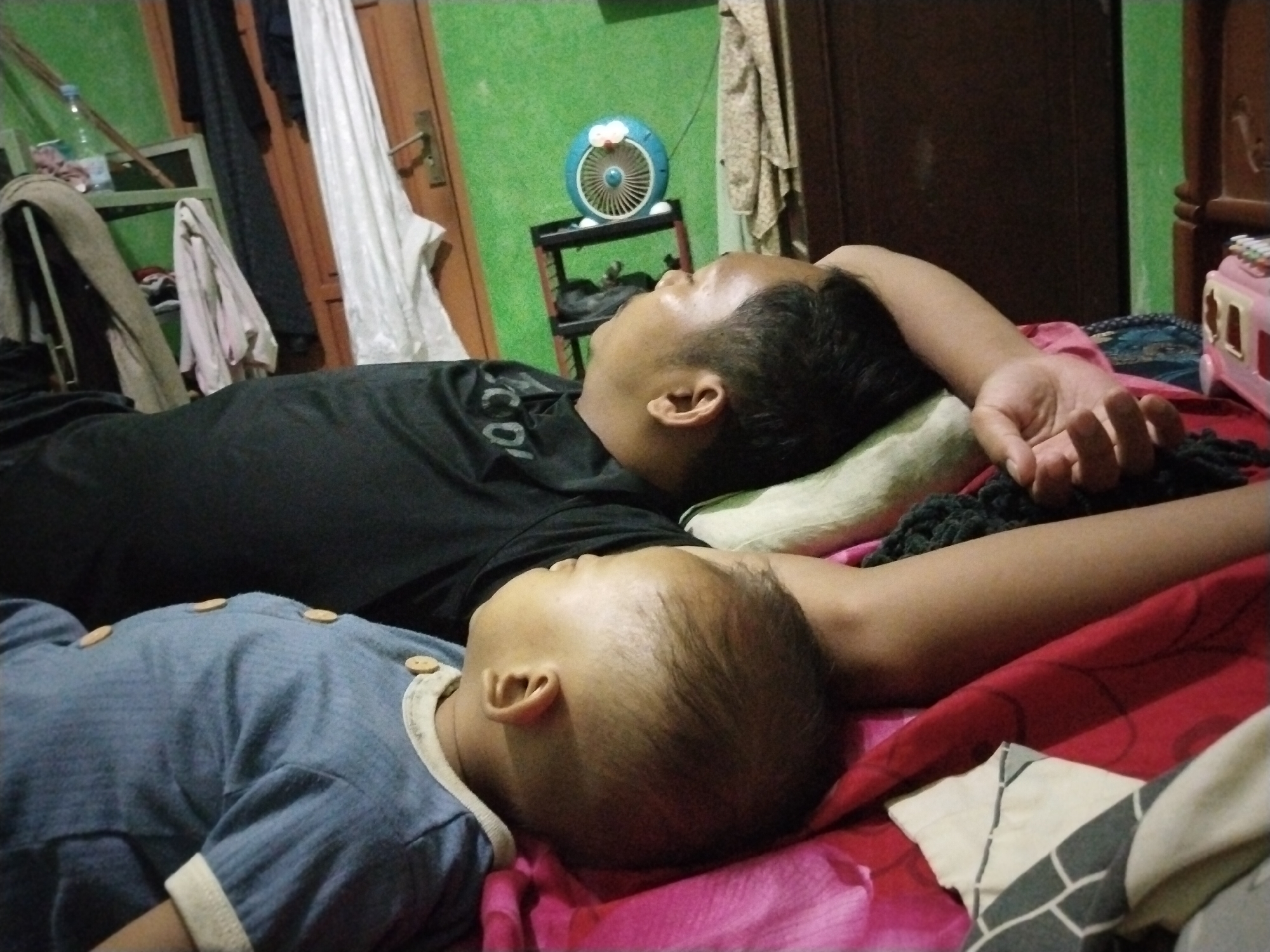 In Kecamatan Pati, Indonesia, an indoor scene captures two individuals resting in close proximity, possibly a family. A man, lying on his back, faces to the right of the frame, his head resting on a pillow. He wears a black t-shirt. Next to him, a baby is also lying down, facing upwards, wearing a blue onesie with light-colored trim and wooden buttons. The background reveals a green painted wall, a brown wooden door, and a shelf unit holding a blue, cartoon-themed fan. Various clothing items and fabrics are visible, including a white garment draped near the door, and what appears to be a pink blanket or sheet beneath the individuals. The overall impression is one of a domestic setting.