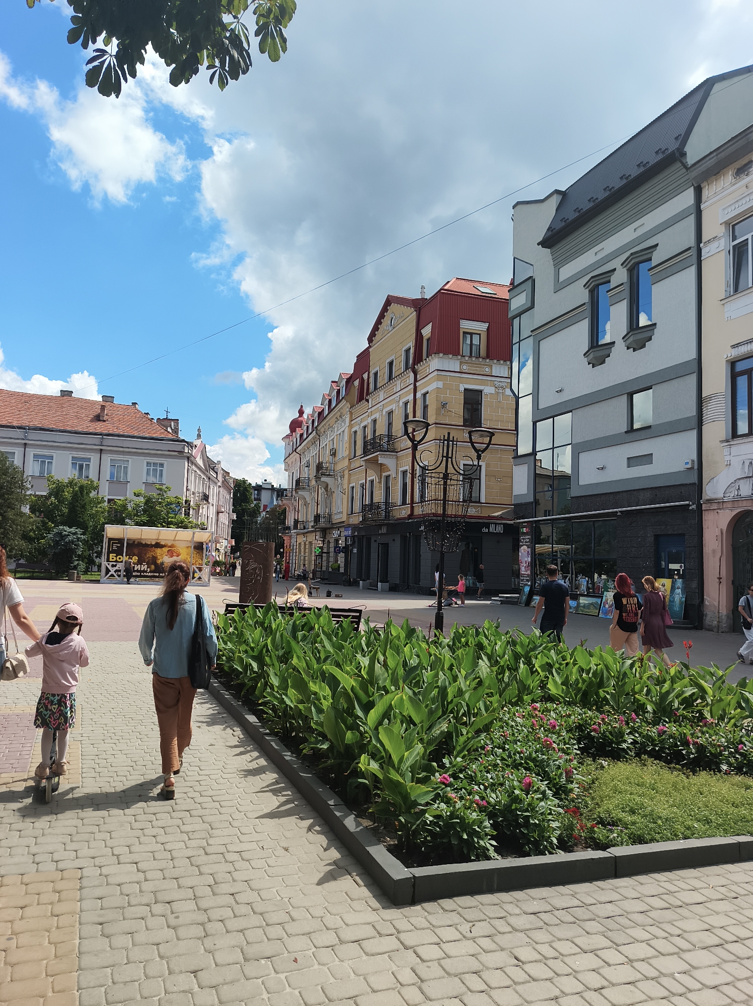The image depicts a sunny day in the city of Ternopil, Ukraine, likely on a pedestrian walkway. In the foreground, a woman and a young child on a scooter are walking away from the camera. The child is wearing a pink hoodie and skirt, while the woman is in a denim shirt and brown pants. To the right of the walkway, there is a raised flower bed with lush green foliage and pink flowers. In the background, various buildings line the street, displaying a mix of architectural styles. One building is particularly tall with a modern glass facade, reflecting the sky. Another building to its left has a distinct yellow exterior with a red roof. Further down the street, more buildings with traditional facades and balconies are visible. People are seen walking along the street and seated on benches. The sky is partly cloudy, with a mix of bright blue sky and white clouds. The overall scene suggests a vibrant urban environment on a pleasant day.