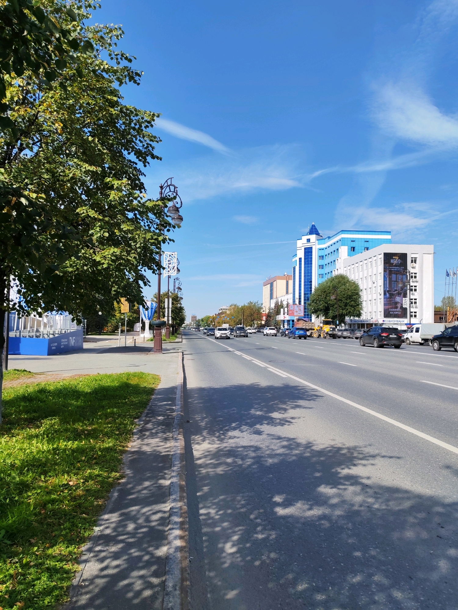 The image depicts a sunny day in Tyumen, Russia, with a wide asphalt road in the foreground and a clear blue sky with wispy clouds above. A tree with green leaves casts shadows on the sidewalk and the road. On the left side of the road, there is a sidewalk, a patch of green grass, and a blue structure with white lettering. Beyond the sidewalk, there are streetlights and a row of buildings. Several cars are visible on the road, and some buildings are in the background, including a modern multi-story building with blue accents. The overall scene is that of a typical urban environment on a bright day.