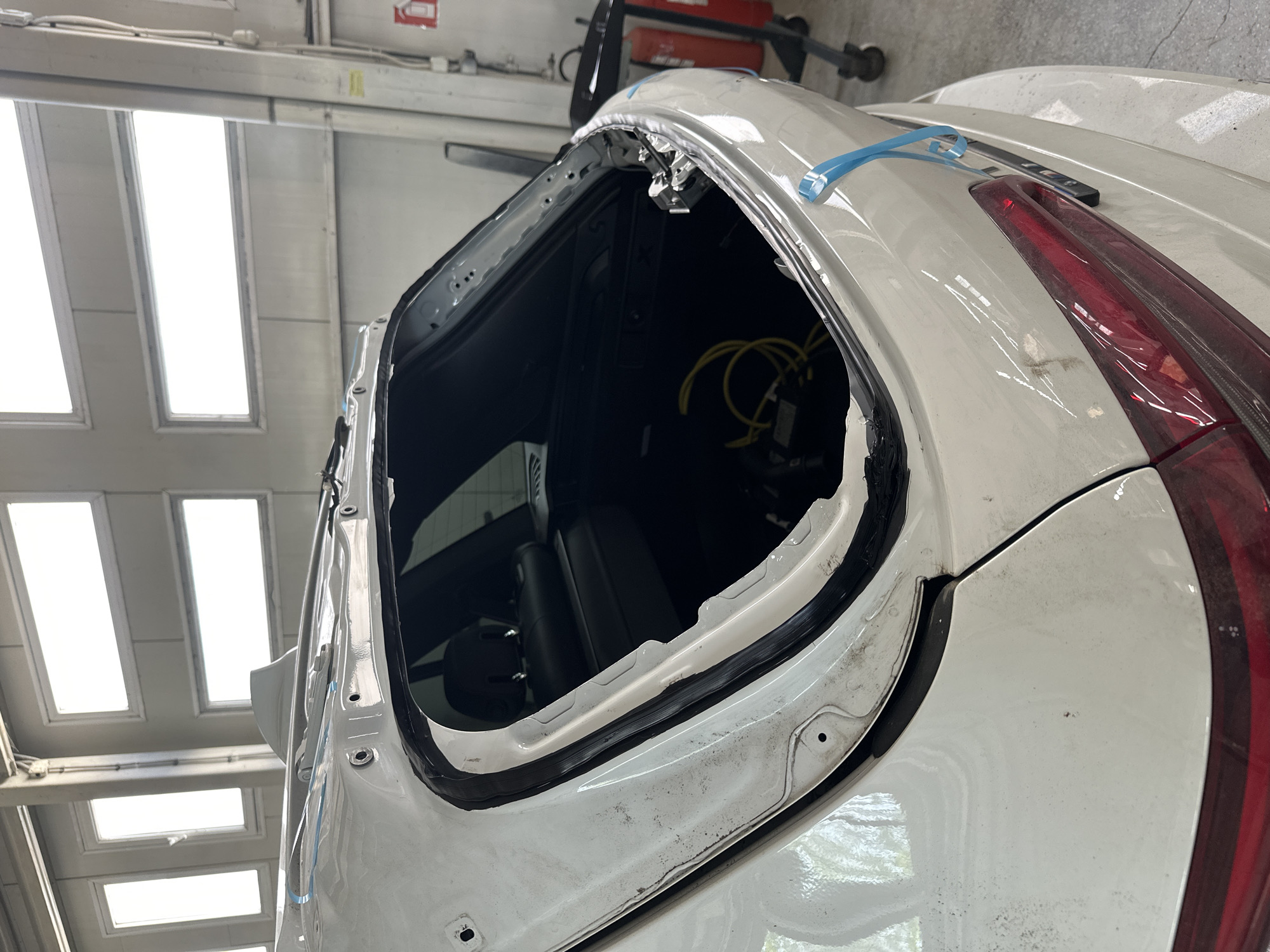 An angled, medium shot captures the rear section of a white car undergoing repair in an indoor automotive workshop. The main subject is the car, specifically its rear window or hatch area, which is currently open or has had its glass removed. A thick, dark, possibly old sealant or adhesive residue is visible along the exposed frame where the window would typically be seated. The white paint of the car shows some dirt and dust, particularly around the open area and on the lower part of the bodywork.
Inside the exposed rear cavity of the car, yellow cables are coiled and visible against the dark interior. A portion of a red taillight is seen on the far right side of the image. Blue masking tape is applied to the car's body panels near the rear window opening, suggesting ongoing work or preparation for a repair or reinstallation.
The setting is a well-lit workshop, with several bright, rectangular overhead lights or skylights visible on the ceiling, casting a strong, even illumination over the scene. The background reveals parts of a grey wall and other indistinguishable equipment typically found in a repair facility. The image is captured from a slightly elevated perspective, looking down into the open section of the vehicle. No discernible text is visible on any objects.