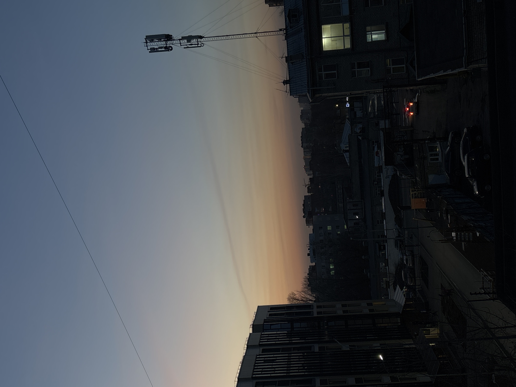 A twilight view from a high vantage point, likely a building, overlooking a cityscape. In the foreground, modern residential or commercial buildings with dark windows are visible. Further back, a dense urban landscape extends into the distance, punctuated by the silhouettes of buildings, trees, and infrastructure. A construction crane is visible at the top of the frame. Cars are parked alongside buildings to the right. A single illuminated window on one of the adjacent buildings provides a stark contrast to the dimness. The sky transitions from a deep blue to a lighter orange and yellow hue at the horizon, suggesting sunset or sunrise. A powerline runs across the sky. Based on the architectural styles, urban density, and presence of the crane and powerlines, it is plausible that the location is Perm, Russia. Specific details regarding people, actions, and interactions are not visible in this still image.