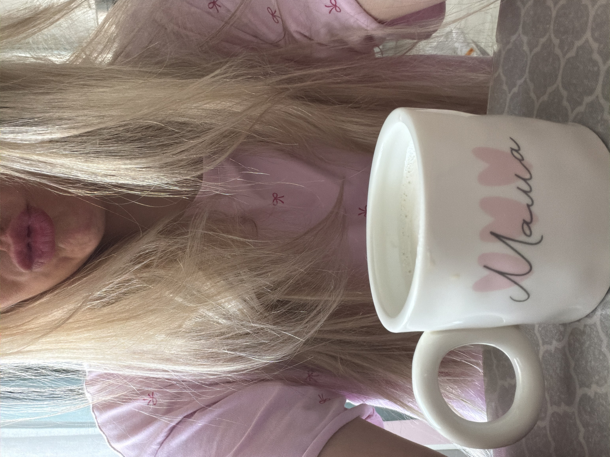 The image, taken in Votkinsk, Russia, captures a close-up view of a person with very long, straight, blonde hair, cascading across much of the frame. The person's lips, coated with a light pink lipstick, are visible in the upper left quadrant. They are wearing a pink garment, possibly pajamas, adorned with small, darker pink bow patterns.
To the right, a white ceramic mug, filled with a light-colored liquid (likely milk or a milky drink), is prominently displayed. The mug features the word "Мама" (Mama), meaning "Mom" in Russian, written in elegant black cursive script, accompanied by several soft pink heart shapes. The mug rests on a gray surface with a subtle geometric or floral pattern, which appears to be a tablecloth or placemat. The scene suggests a relaxed, indoor setting, likely during the daytime, conveying a calm and personal moment.