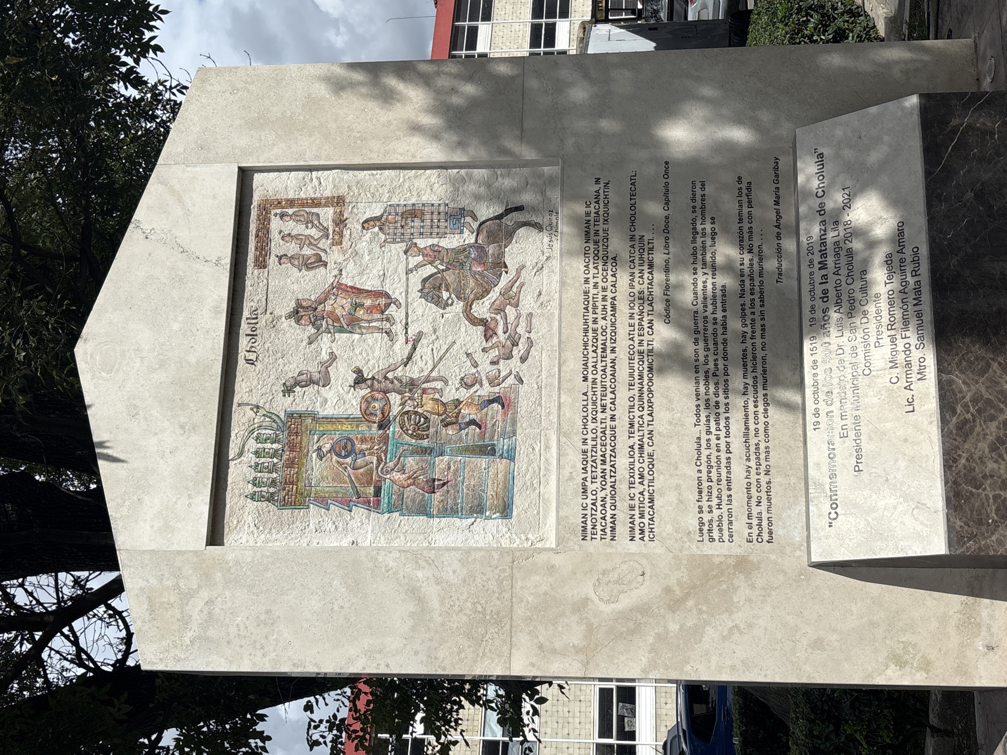 A vertical stone monument stands outdoors in San Pedro Cholula, Mexico, against a backdrop of dense tree foliage and a multi-story building. The monument consists of a light-colored rectangular slab, topped with a peaked cap.
Centered on the monument is a vibrant, rectangular mosaic depicting historical events. The top-left of the mosaic is inscribed with "Chololla." The mosaic illustrates a tumultuous scene featuring numerous figures: indigenous people, characterized by dark skin, traditional attire including loincloths, skirts, and feathered headdresses, some carrying round shields and spears or clubs; and Spanish conquistadors, depicted with lighter skin, wearing metal armor, with one figure on a brown horse, wielding a sword, trampling over fallen indigenous figures. Other indigenous figures are shown seated, standing, or engaged in conflict. Indigenous architectural elements, possibly temples or palaces, are visible in the upper portion of the mosaic. The artist's name, "Jesús Quiroz Chimalp" is faintly visible below the mosaic on the right side.
Below the mosaic, two blocks of text are inscribed. The upper block is in Nahuatl, beginning with "NIMAN IC UMPA IAQUE IN CHOLOLLA..." The lower block provides a Spanish translation, detailing the arrival in Cholula, a gathering in the "patio de dios," and subsequent "acuchillamiento, muertes, golpes." It mentions the Cholulans' lack of fear and their death without resistance or knowledge. This text concludes with the citation "Códice Florentino, Libro Doce, Capítulo Once."
Affixed to the lower front of the monument is a darker, polished stone plaque. This plaque bears the dates "19 de octubre de 1519" and "19 de octubre de 2019," marking the "Conmemoración de los 500 años de la Matanza de Cholula" (Commemoration of the 500 years of the Cholula Massacre). It further lists "En mandato de Dr. Luis Alberto Arriaga Lila, Presidente Municipal de San Pedro Cholula 2018 - 2021" and names from the "Comisión De Cultura": "Presidente C. Miguel Romero Tejeda, Lic. Armando Filemón Aguirre Amaro, Mtro. Samuel Mata Rubio."
The monument is positioned in an urban environment, with the dense green leaves of a large tree visible behind its upper left side, and a modern building with multiple windows in the background on the right.