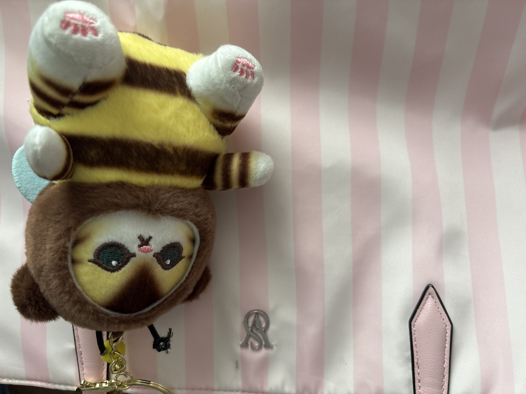 The image shows a plush toy hanging upside down on a pink and white striped bag. The toy features a brown, round face with large eyes and a small pink nose. Attached to the face is a yellow and brown striped body with white paws that have pink paw prints. A small light blue wing is visible on the side of the body. The bag has a silver "AS" logo and a pointed pink tab. The location is unknown.