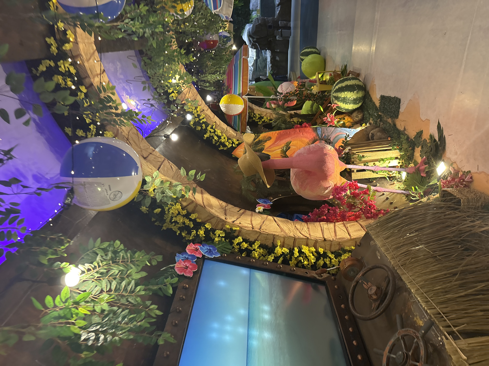 The image depicts an indoor entertainment or themed area in West Jakarta, Indonesia, designed with a tropical beach motif. No individuals are present in the scene.
In the foreground on the left, a rustic, low-lying structure is present, clad in a light brown, frayed, straw-like material. On its flat top surface, two weathered, multi-spoked ship's wheels are mounted, alongside a small, rust-colored lever. To the immediate right of this structure, a large, pink flamingo statue stands prominently, adorned with a straw sun hat. Its base is surrounded by artificial red and pink flowers, and green foliage.
Behind the foreground structure, a large rectangular screen displays a tranquil image of a clear blue sky meeting an ocean horizon, with gentle waves. The screen is framed by a dark, metallic border secured with rivets. Artificial red and blue hibiscus flowers are strategically placed on the screen's frame. Adjacent to the screen and arching into the background, a curved architectural element is decorated with garlands of small yellow artificial flowers and green vines.
The ceiling above is a mix of brown textured surfaces and areas illuminated by purple uplighting. Various artificial green vines hang down, interspersed with warm white string lights and exposed bare bulb lights. Large blue and white beach balls are suspended from the ceiling in multiple locations.
Towards the center and right mid-ground, a vibrant tropical setting unfolds. Several oversized, round props resembling watermelons (some striped green, others solid lime green) are arranged on the light-colored, smooth floor, which features irregular orange-brown patches resembling sand or dirt. Another pink flamingo statue is partially visible behind the first, and a tall, orange and yellow patterned surfboard prop stands upright. A small table with yellow beverage cups is also visible. An open beach umbrella with red, orange, yellow, green, and blue stripes provides a central focal point. In the background, rock formations and a water feature or pool are visible, suggesting an aquatic element to the environment. Further string lights and green foliage enhance the tropical ambiance throughout the upper areas of the scene.