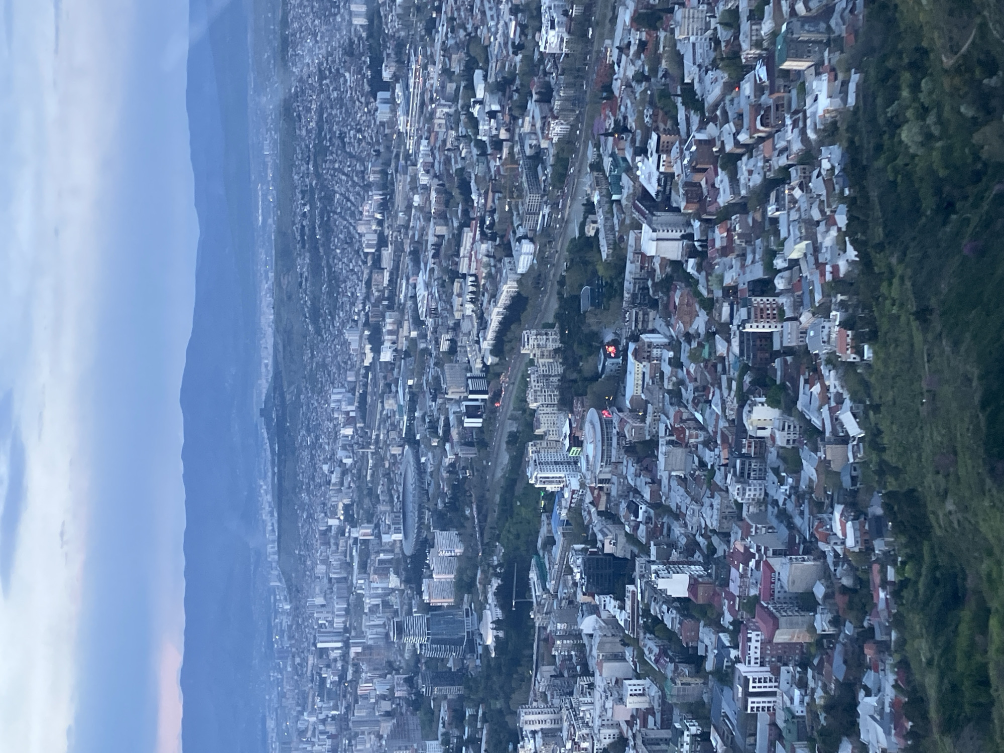 An aerial, slightly rotated view captures the sprawling city of Tbilisi, Georgia, under the soft light of dusk or dawn. The image, if rotated 90 degrees counter-clockwise, would show the city extending into the foreground, bordered by a dense, forested hillside on the right. In the distance, a range of mountains rises, their peaks partially obscured by haze.
The urban landscape is dense with numerous buildings of varying sizes, exhibiting a mix of residential and commercial structures. A prominent linear green space, possibly a river or park, winds through the city, dividing different neighborhoods. Several city lights are visible, indicating the low light conditions of the early morning or late evening. A large, distinctive circular building, possibly a stadium or cultural center, stands out within the urban fabric.
No specific human activity is discernible from this high vantage point. The sky is largely overcast or hazy, contributing to the muted color palette of blues and grays, with subtle hints of warmer tones near the horizon. There is no visible text in the image.