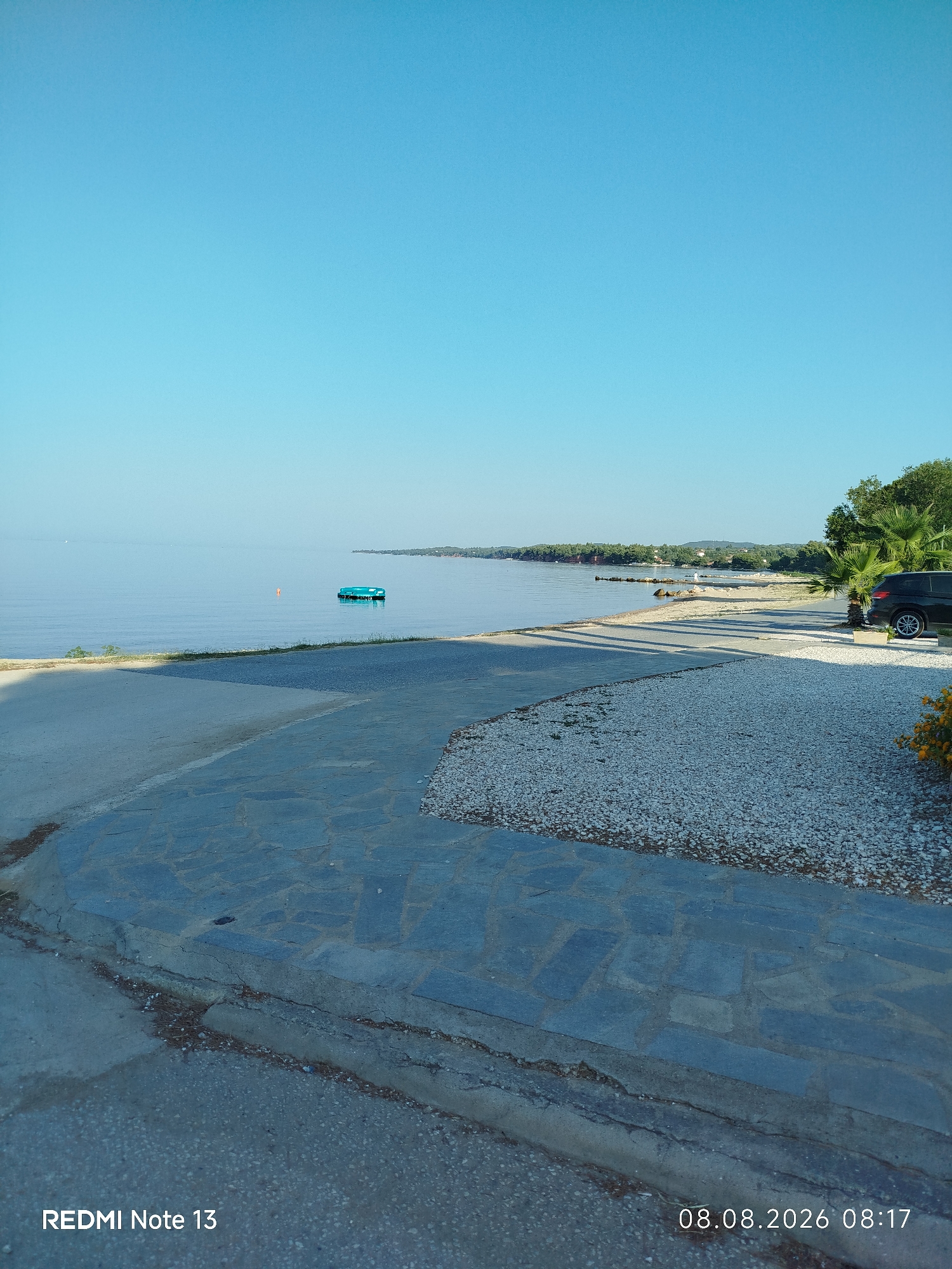 The image depicts a tranquil seaside scene on a clear day, likely in Sithonia Municipal Unit, Greece. In the foreground, a paved area transitions from concrete to irregularly shaped stone tiles, bordered by gravel. To the left, the calm, blue water of the sea stretches towards the horizon. A small, bright green floating structure, possibly a buoy or a small raft, is visible on the water. Further in the distance, a treelined shore with a sandy beach is discernible, punctuated by scattered rocks. On the right side of the frame, a dark-colored SUV is parked on a gravel surface near palm trees. A patch of vibrant orange flowers adds a splash of color to the lower right corner. The sky is a uniform, clear blue. The image is dated August 8, 2026, at 08:17, as indicated by the timestamp at the bottom right. The text "REDMI Note 13" is present at the bottom left.