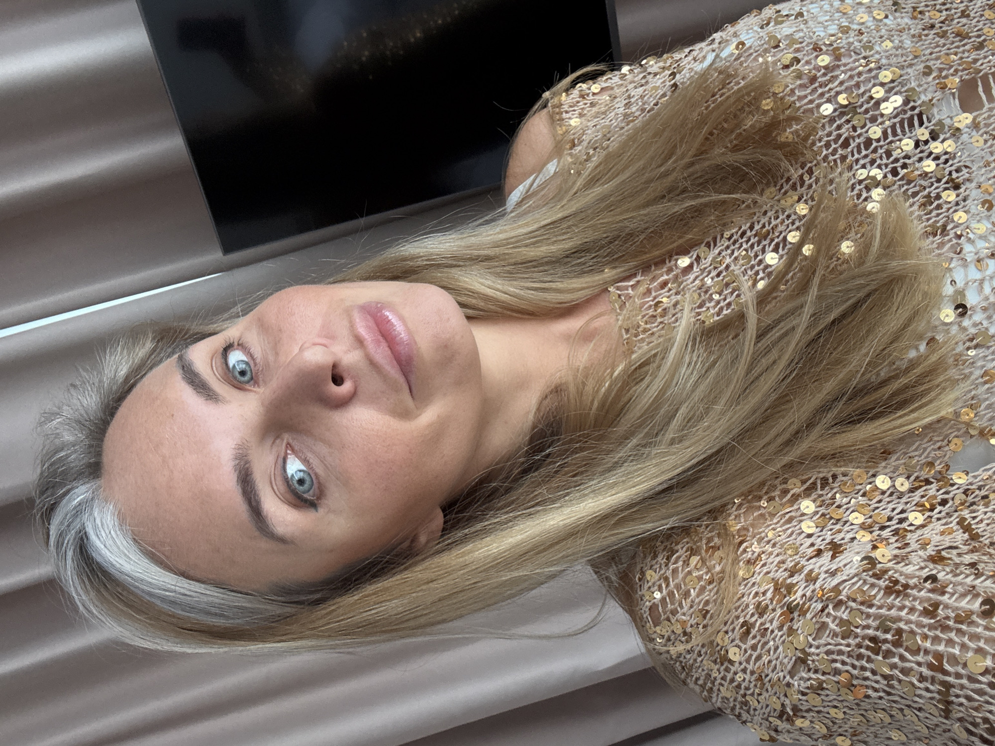 A woman with blonde hair, some gray streaks, and blue eyes looks into the camera. She is wearing a shimmering gold sequined sweater. The background consists of draped curtains and a dark television screen. The image is a selfie taken from the chest up. The location is the city of Wuxing District, China.