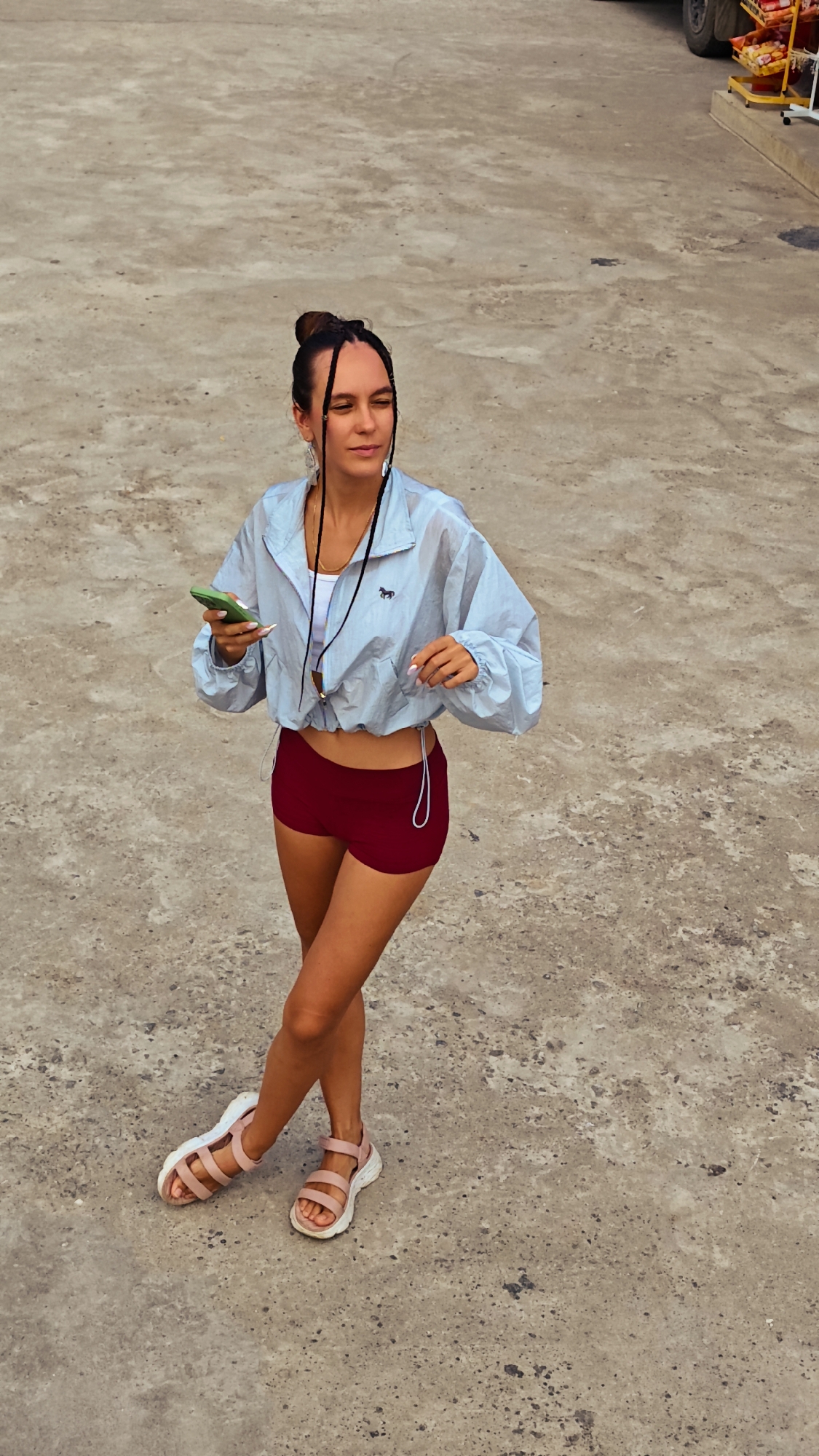 A young woman stands outdoors on a concrete surface. She has dark hair pulled up in a bun with thin braids framing her face. She is wearing a light blue jacket with a small horse logo, a white top, and burgundy shorts. She is also wearing pink sandals with white soles. She holds a green smartphone in her left hand and has white earphones hanging around her neck. Her legs are crossed at the ankles.
Behind her, on the right side of the frame, there is a vehicle with a cart attached, possibly a fruit stand.
The location is implied to be within Lũng Phìn Commune, Vietnam.