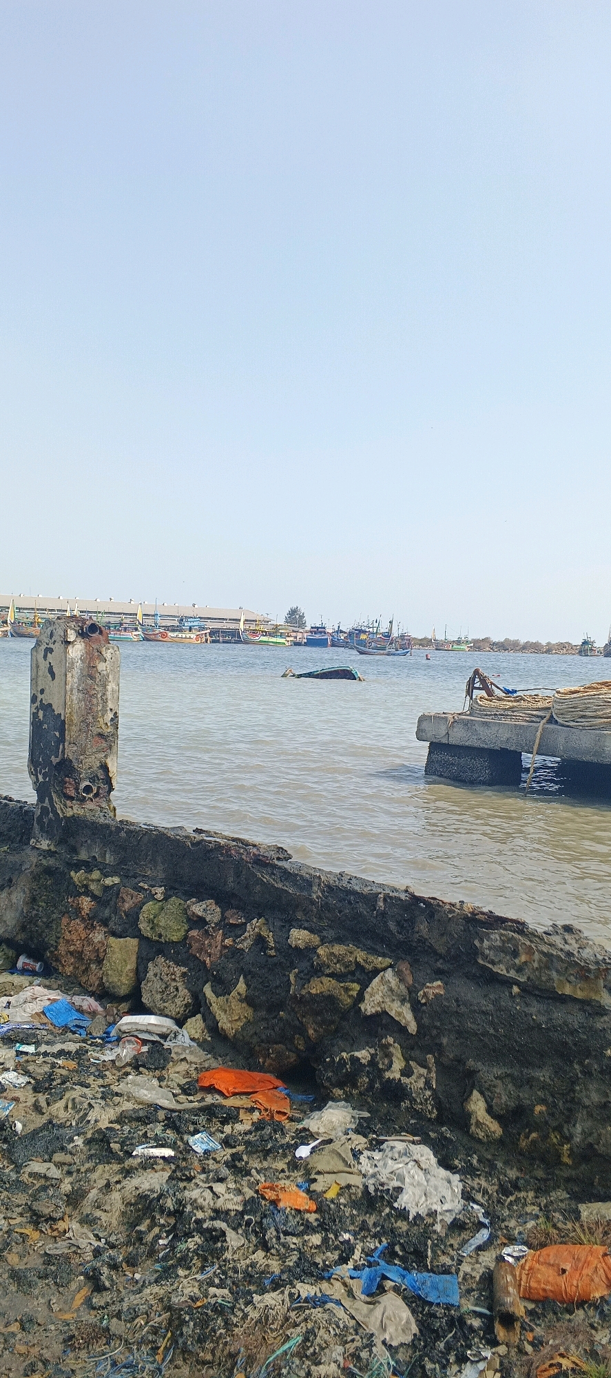 The image depicts a polluted waterfront scene in Blimbing, Indonesia. In the foreground, a pile of debris, including plastic bags, bottles, and other refuse, is accumulated against a decaying concrete and stone structure. A rusted metal pipe protrudes from the structure. The water is murky and appears to be a bay or harbor, with a concrete pier visible on the right side, laden with coiled ropes. In the middle ground, a partially submerged object, possibly a capsized boat, is visible. The background features a line of various fishing boats moored near buildings that suggest a port or market area. The sky is clear and blue, indicating a daytime scene. There are no people visible in the image.