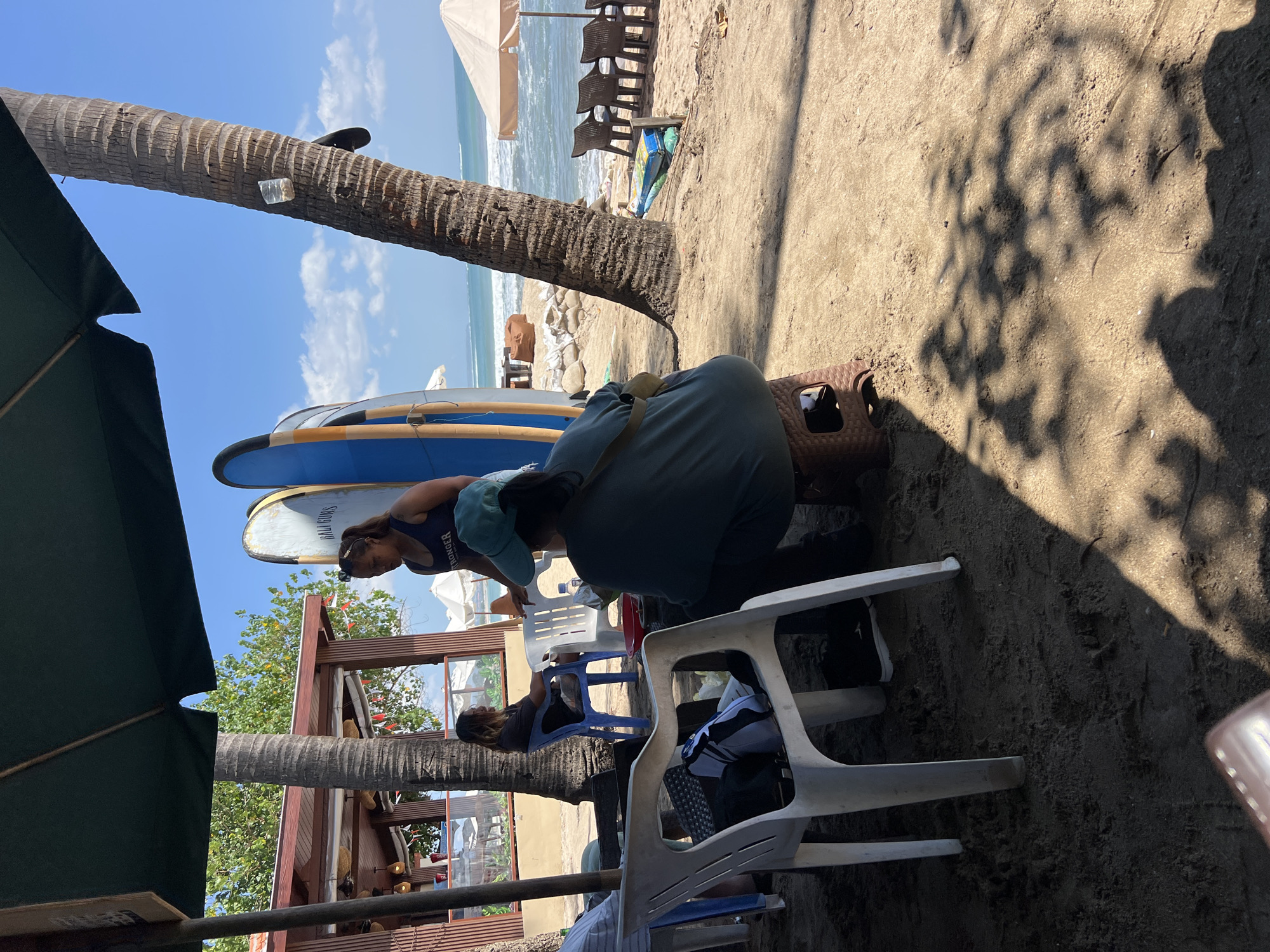 The image shows a beach scene in Kuta, Indonesia, likely during the day. In the foreground, two white plastic chairs are visible on the sand, one with a black and white bag on it. A person is sitting on a brown stool to the right, facing away from the camera, wearing a dark blue top and a teal hat. Another person, wearing a dark blue tank top with text that appears to say "STRONGER," is standing and interacting with the seated person. Behind them, two surfboards stand upright, one blue and one off-white with text. A palm tree trunk is prominent to the right of the surfboards. In the background, a section of a wooden structure, possibly a restaurant or bar, is visible, with tables and chairs set up. Further back, umbrellas and chairs are arranged on the sand, and the ocean with moderate waves is visible under a clear blue sky with scattered clouds. The overall scene depicts a relaxed atmosphere on a tropical beach.