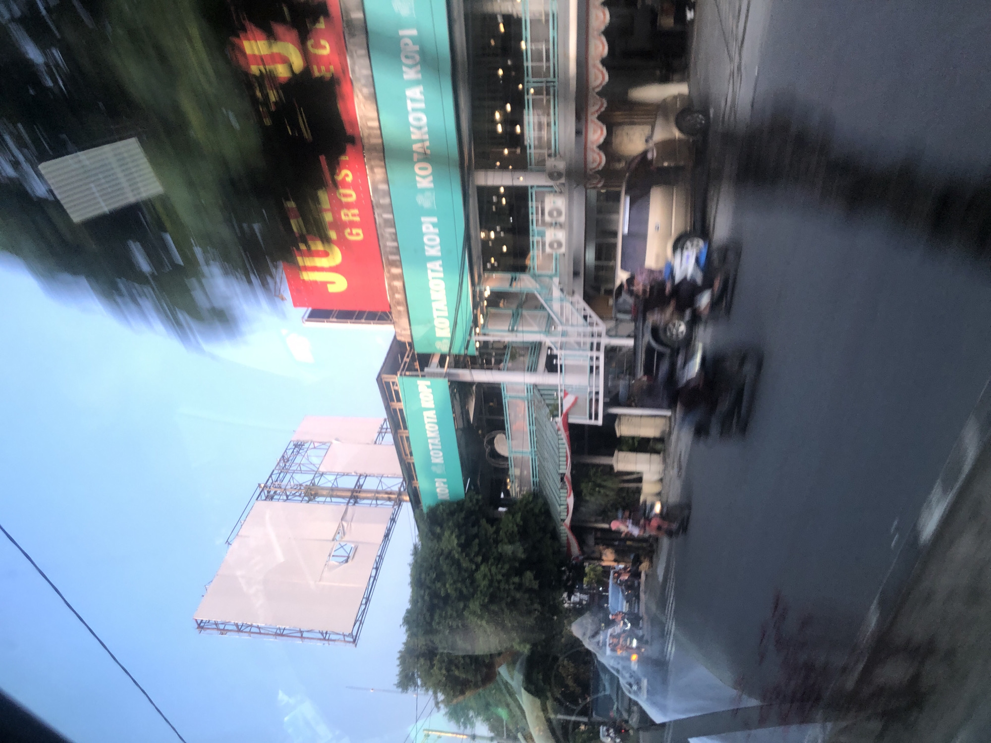 The image shows a street scene in Medan, Indonesia, likely on Jalan Gatot Subroto, based on the presence of a "Kotakota Kopi" establishment. The foreground is dominated by a blurred road and pavement, with several motorcycles and a few cars in motion, indicating a busy urban environment. A light-colored van is visible on the right side of the road.
In the background, a multi-story building houses a "Kotakota Kopi" cafe. The building has a distinctive teal-colored awning with "KOTAKOTA KOPI" written in white lettering. Above this, a red billboard with yellow lettering, partially obscured by trees and motion blur, appears to advertise "JUAL GROSIR" (Wholesale Sale). A large, white billboard structure is also visible in the upper left portion of the image. Trees and greenery are present, suggesting a somewhat developed urban area with some vegetation. The sky is a clear blue, indicating daylight. The overall impression is a dynamic streetscape in a commercial district.