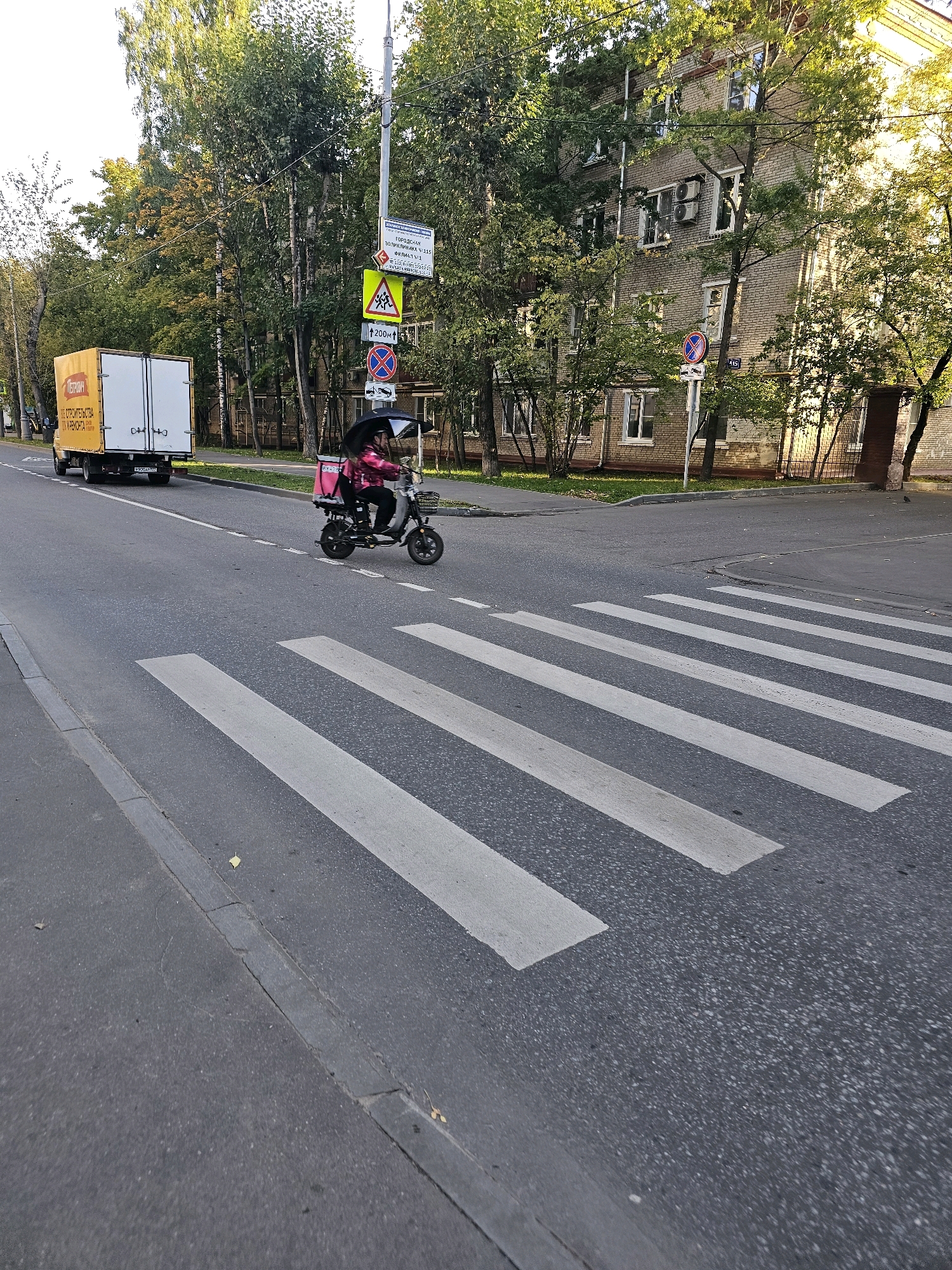 A sunny daytime exterior scene in the city of Khimki, Russia, features a street with a pedestrian crossing prominently in the foreground. On the road, a person is riding a three-wheeled electric scooter, holding an umbrella for shade. The scooter is pink and has a black storage box on the back, suggesting it might be used for deliveries. In the background, a yellow delivery truck with white doors is parked on the side of the road. Behind the truck and scooter, a multi-story brick apartment building is visible, with trees lining the street and sidewalk. Traffic signs are also present on a pole near the apartment building, including a pedestrian crossing warning sign and a circular sign with a red border and a blue center. The street is paved, and a curb separates the road from a grassy area and sidewalk.