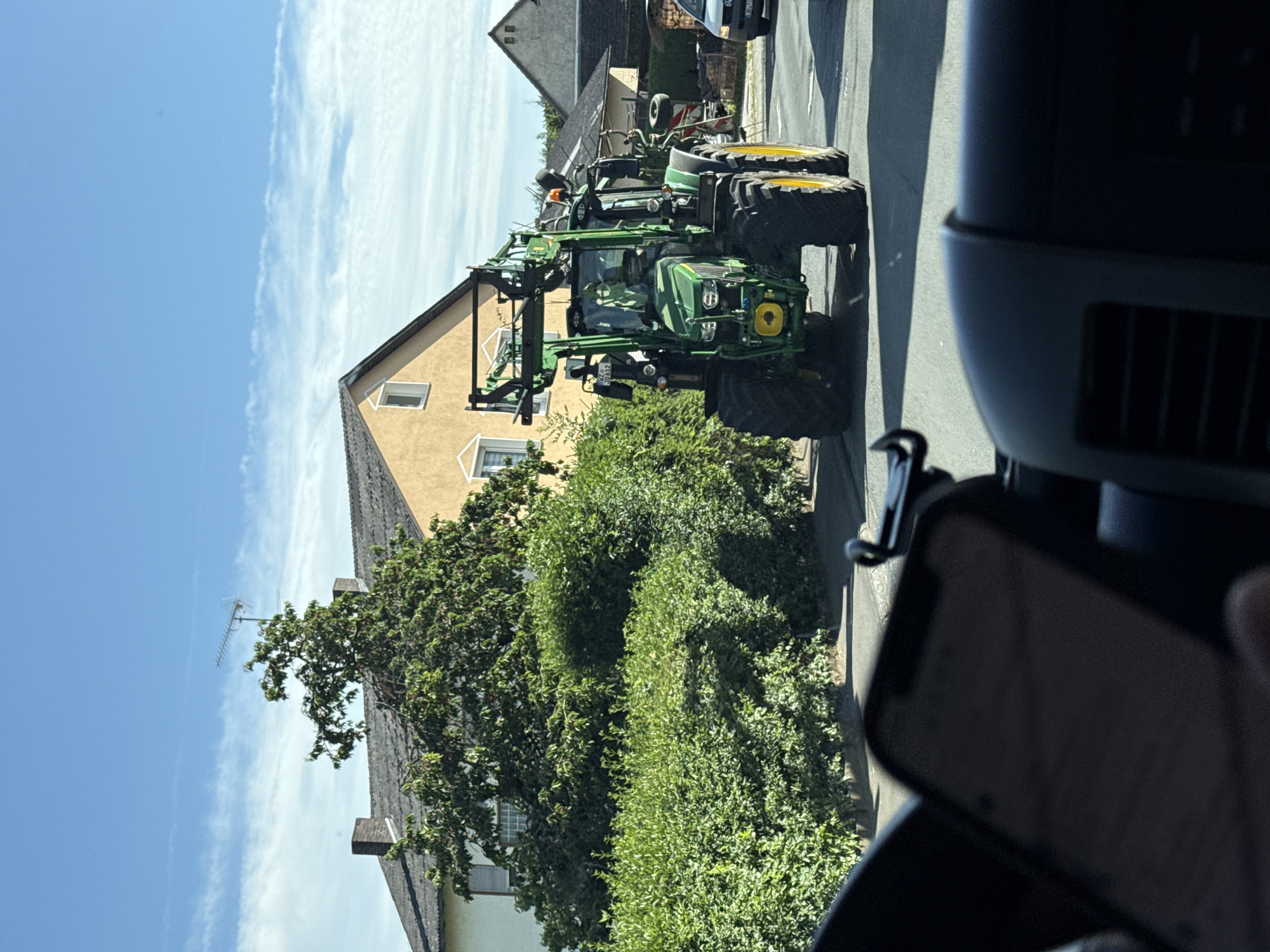The image depicts a green John Deere tractor with yellow wheels driving down a street in Volpertshausen, Germany. The tractor is equipped with a front-end loader. On the left side of the street, a lush green hedge runs along the property line of a yellow house with a gray tiled roof. A mature tree with dense foliage is situated in front of the house. The sky is blue with scattered white clouds. In the foreground, the interior of a vehicle is visible, with a smartphone mounted on a holder. Another vehicle is partially visible in the background on the right side of the road.