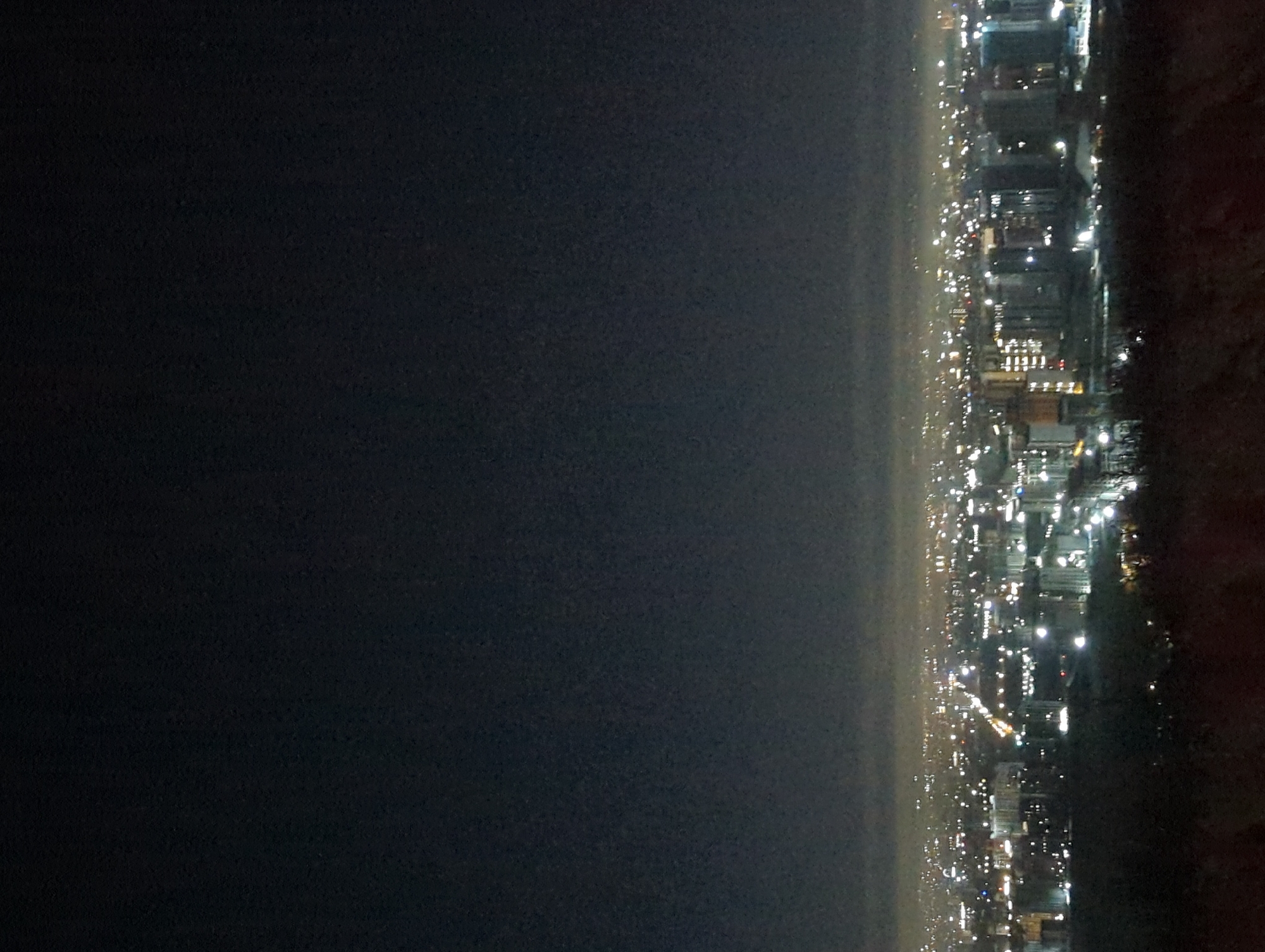 The image shows a nighttime view of Bishkek City, Kyrgyzstan. The right side of the image depicts a line of illuminated buildings and streetlights stretching into the distance, suggesting a city skyline along a coast or border. The left side of the image is dark, likely representing a body of water or open space.