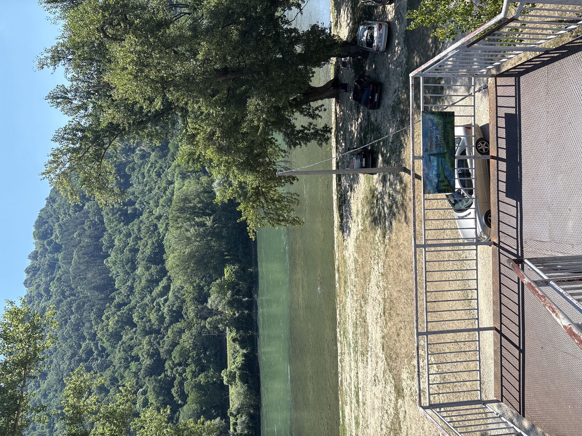 The image depicts a serene riverside landscape on a clear, sunny day. The foreground features a metal staircase leading down from an elevated viewpoint, with a railing overlooking a sandy embankment. Beyond the embankment, a wide, calm river with green, clear water flows, bordered by lush green trees and a forested hillside in the background. Several cars are parked on the sandy area near the riverbank, under the shade of a large tree. A banner with text and a landscape image is attached to the railing in the foreground. The overall scene suggests a recreational area, possibly a park or a picnic spot, situated in a natural setting near Zvornik, Bosnia and Herzegovina.