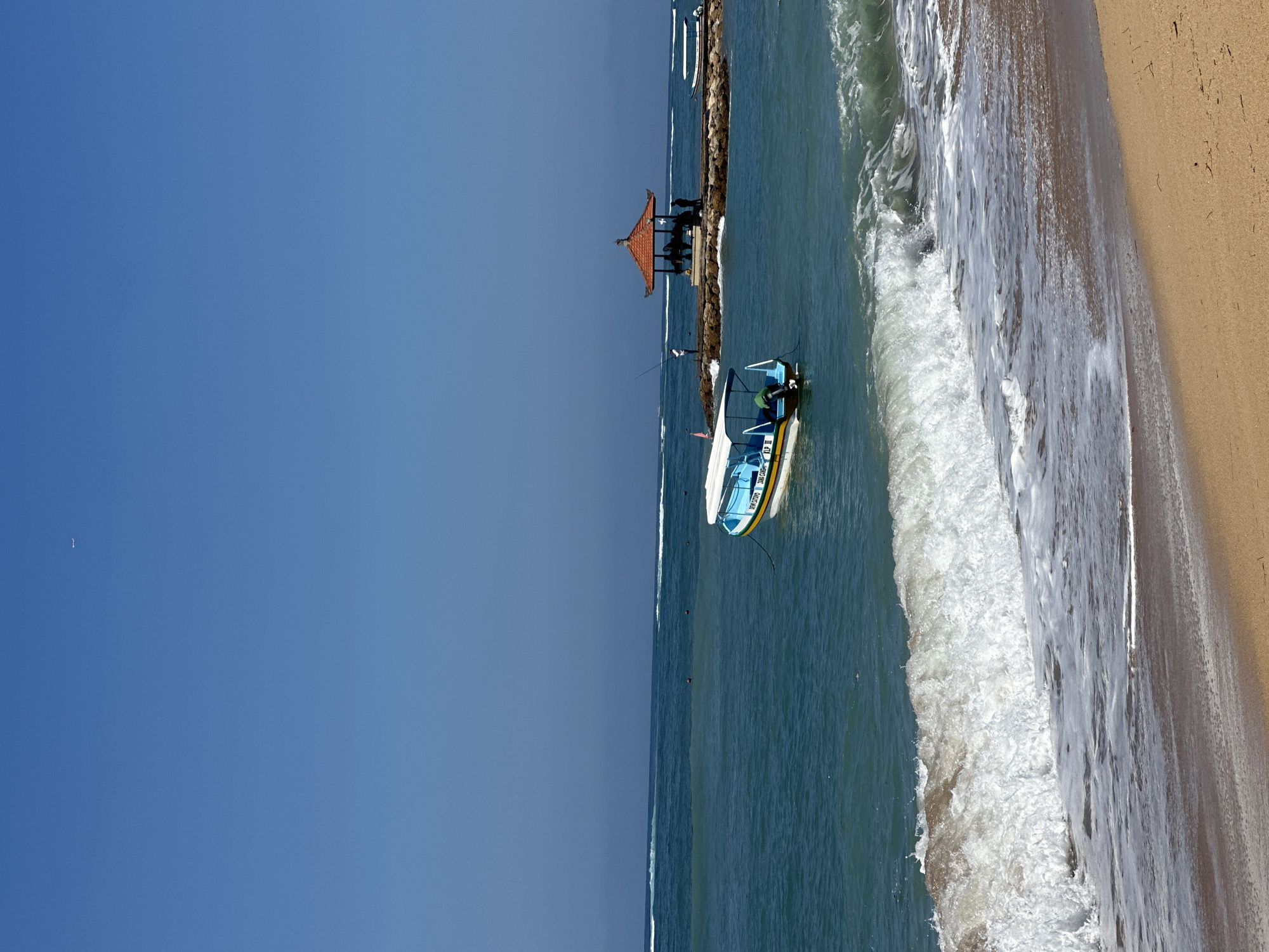 This is a daytime, eye-level, outdoor, full shot of a beach scene in Denpasar, Indonesia. A sandy beach occupies the foreground, with a crashing wave of white foam and turquoise water breaking onto the shore. Beyond the immediate surf, the turquoise ocean stretches to the horizon under a clear, pale blue sky. In the mid-ground, a small, light blue and white motorboat with a yellow stripe is anchored. Further out, a man-made rock pier juts into the water, topped with an octagonal, orange-roofed gazebo. Several people are visible on and around the pier, and another small boat is docked on the far right side of the pier. The sky is mostly clear, with a faint, distant object that may be an airplane visible in the upper left quadrant.