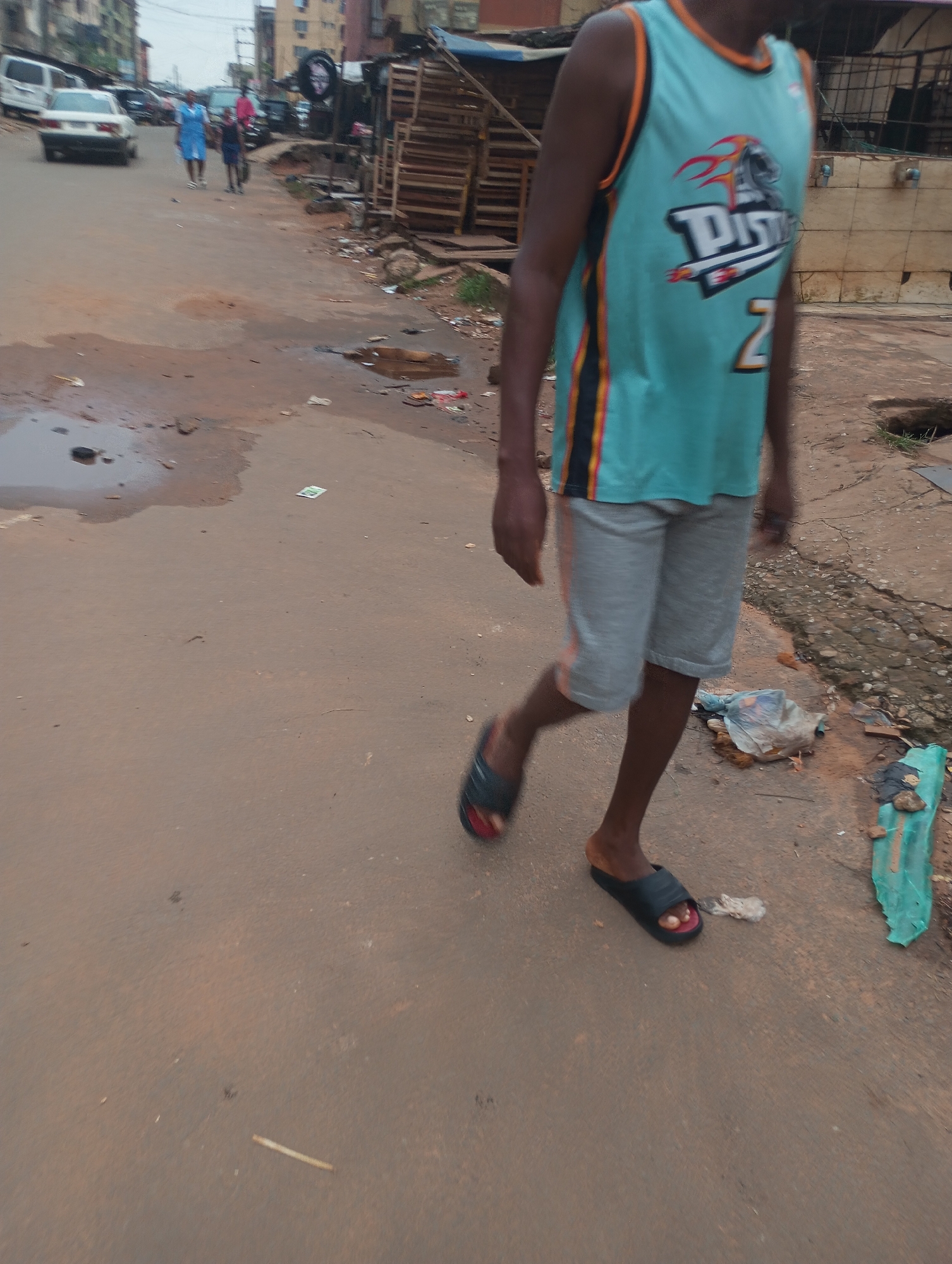 This is a medium shot image taken on a street in Onitsha, Nigeria. The street surface is paved with reddish-brown dirt and has patches of standing water and debris, indicating a potentially neglected urban environment. In the foreground, a person is walking away from the camera, wearing a teal sleeveless jersey with orange and black trim, grey shorts, and black and red slides. The jersey has a graphic design with flames and the word "PISTONS" and the number "2" visible. In the background, several cars are parked or driving on the street. To the right of the frame, there are structures that appear to be informal market stalls or workshops made of wood and corrugated metal, with stacked wooden pallets. Further in the background, there are multi-story buildings, suggesting a densely populated urban area. The lighting suggests an overcast day.