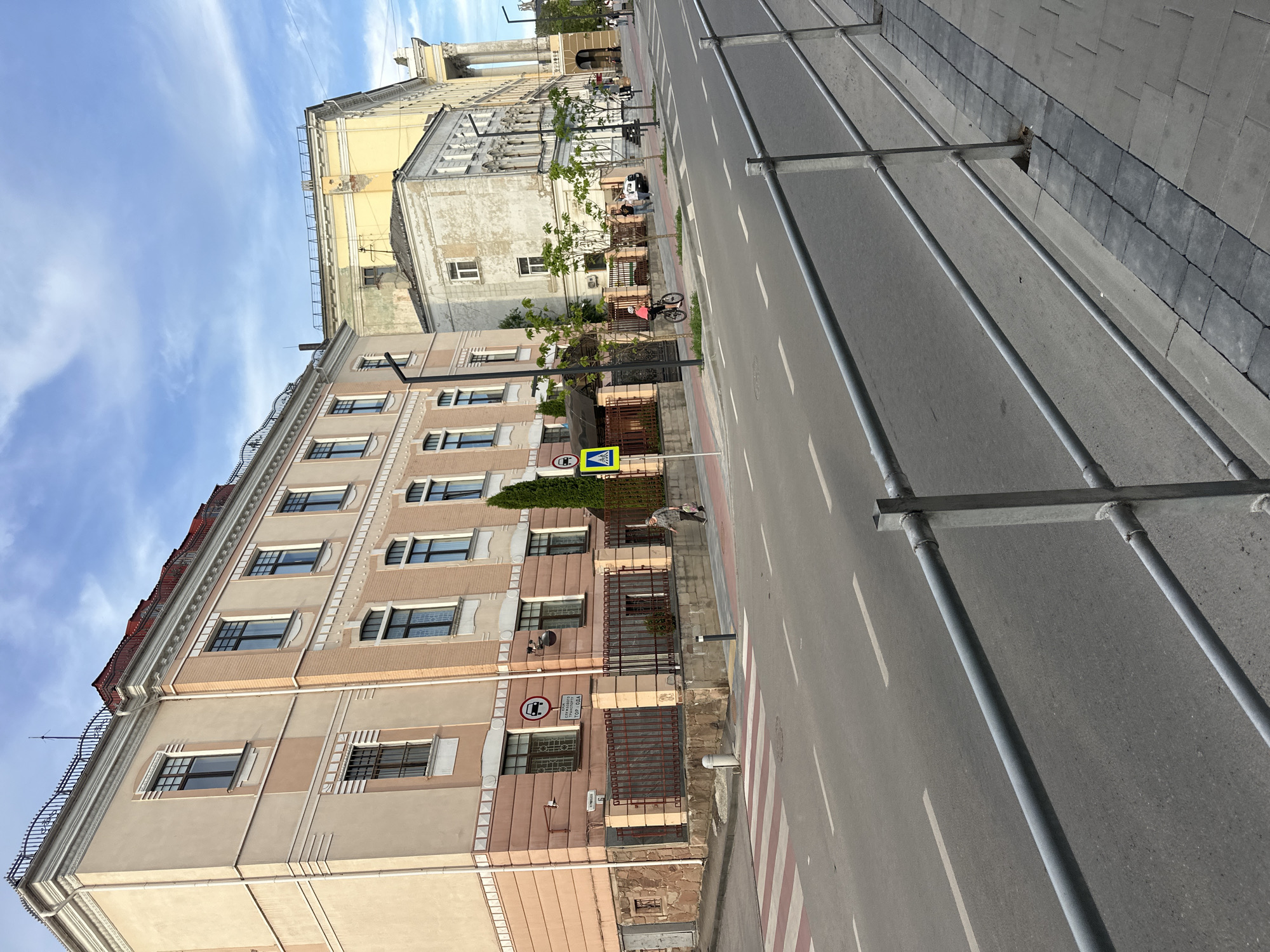 The image depicts a street scene in Ternopil, Ukraine. In the foreground, a grey metal railing runs along a sidewalk paved with grey rectangular stones. Beyond the railing is a street with white lane markings. On the opposite side of the street, a pedestrian crossing with red and white stripes is visible.
Several buildings line the street. The building closest to the foreground on the left is a multi-story structure with a beige facade and pink accents. It features dark-framed windows and decorative cornices. A wrought-iron fence encloses the property in front of this building, and a sign with a car icon and Cyrillic text is mounted on the building.
Further down the street, other buildings are visible, including a yellow building and a lighter-colored, weathered building with arched architectural elements. The sky above is blue with scattered white clouds.
Several people are present in the scene. A woman is walking on the sidewalk in front of the beige and pink building, carrying a bag. A young person on a bicycle is riding on the sidewalk near the pedestrian crossing. Two individuals are standing on the sidewalk further down the street, and other figures are visible in the distance. A black car is parked on the side of the road. Trees and streetlights are also present along the sidewalk.