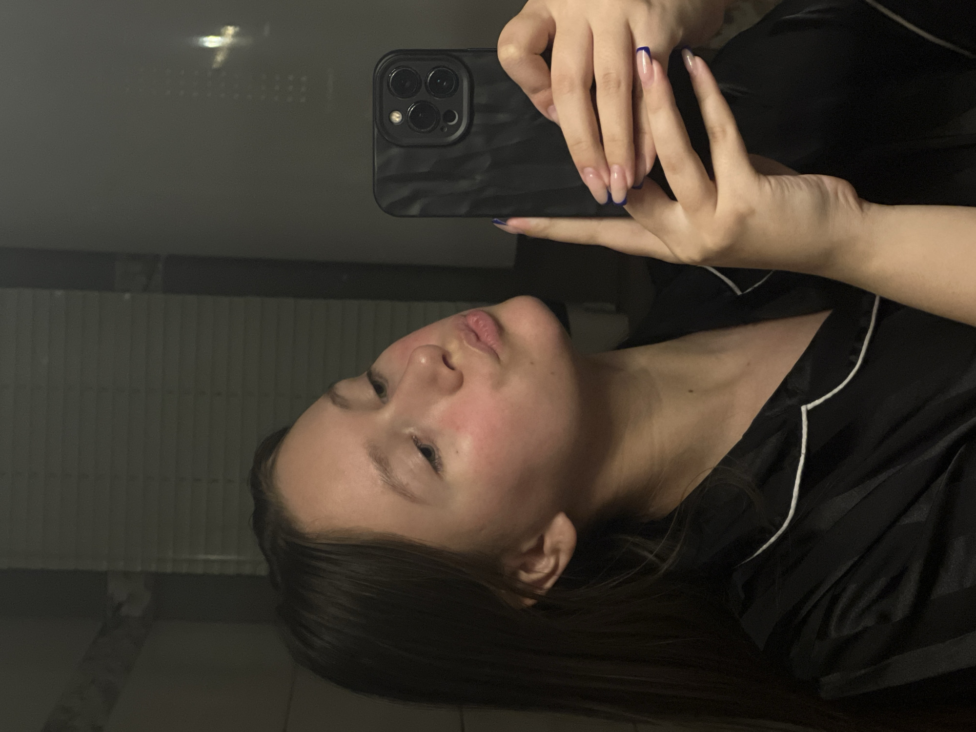 The media file captures a mirror selfie of a young individual, likely female, taken in an indoor setting in Chaikovsky, Russia. The image is rotated 90 degrees clockwise.
The main subject is a young person with fair skin and dark hair, visible from the chest up. They are looking towards their reflection in the mirror, with their lips slightly pursed in a mild pout. There is a noticeable flush or redness on their cheek and jawline. They are wearing dark clothing, possibly satin pajamas, featuring white piping along the edges.
The person holds a dark-colored smartphone with a textured case in their hands, pointing the camera towards the mirror to capture their reflection. The phone features a triple-lens camera system on its back. Their fingernails are styled with a French manicure, characterized by clear or light pink bases and distinct dark blue tips.
The background is dark and appears to be an interior space, likely a bathroom or bedroom, given the presence of a reflective surface. The lighting is dim, suggesting it may be evening or night. No specific location cues beyond the reflective surface are clearly identifiable, though faint patterns or reflections are visible in the darker areas of the image. There is no visible text in the image.