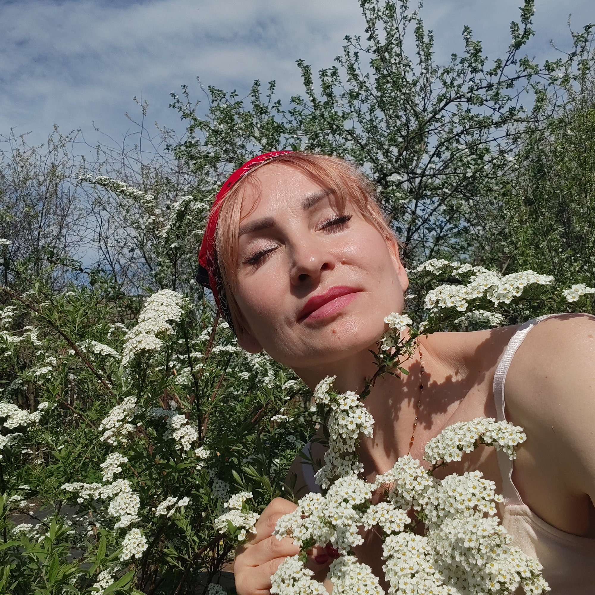 The image features a woman with light pink hair and a red bandana, eyes closed, surrounded by a bush of small white flowers. She is holding a branch of the same flowers near her chest. She is wearing a light-colored top with thin straps. The background includes more flowering bushes and a partly cloudy sky. The location is Chaikovsky, Russia.