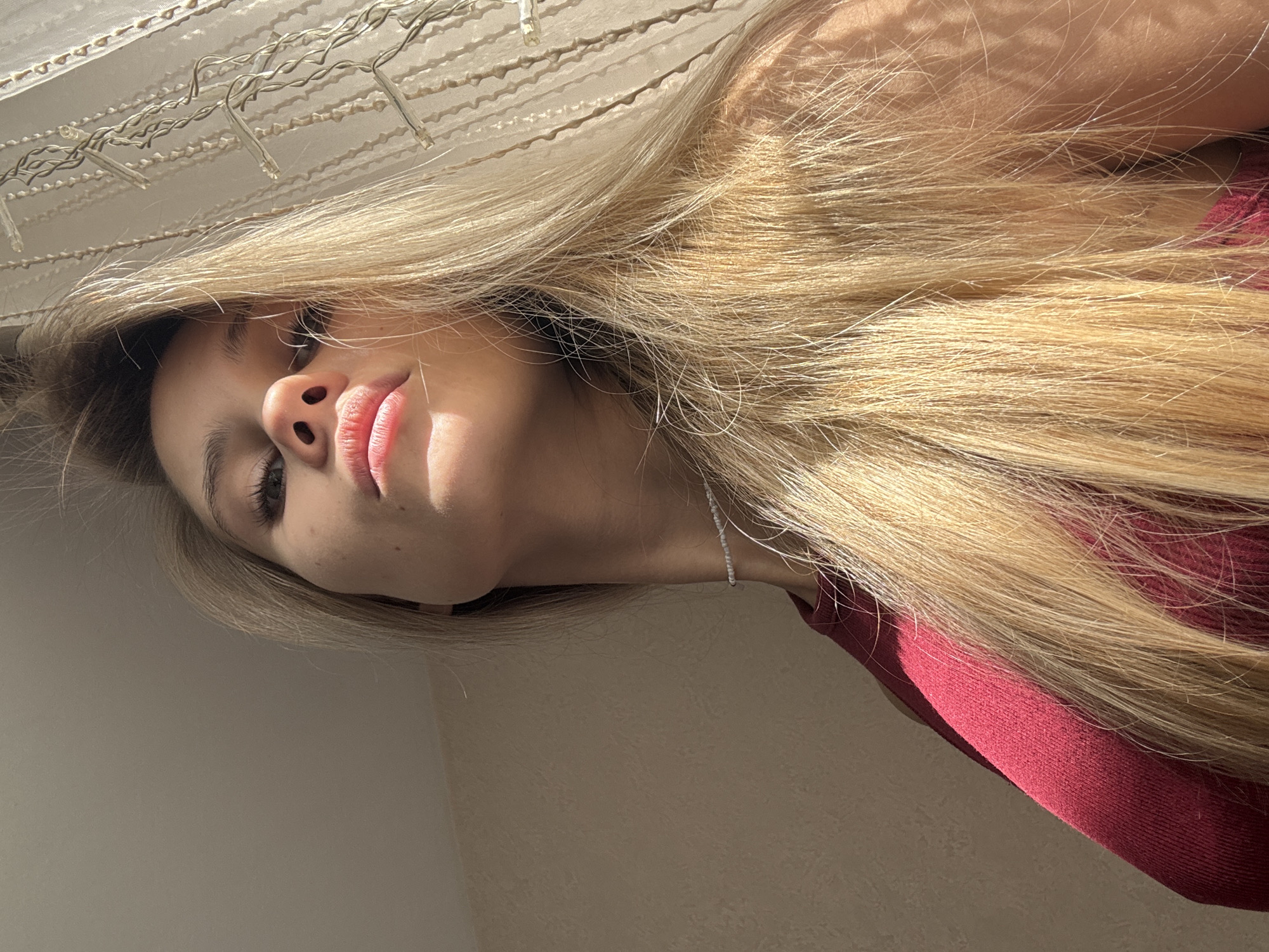 A low-angle, close-up shot shows a young woman with long, blonde hair, wearing a dark red shirt. She is looking up towards the camera, with her face partially in shadow from the sunlight coming from the right. Her lips are parted slightly, and she has a necklace made of small white beads around her neck. Behind her, a sheer white curtain with decorative lights is visible. The background wall is a muted beige color. This image was taken in the city of Chaikovsky, Russia.