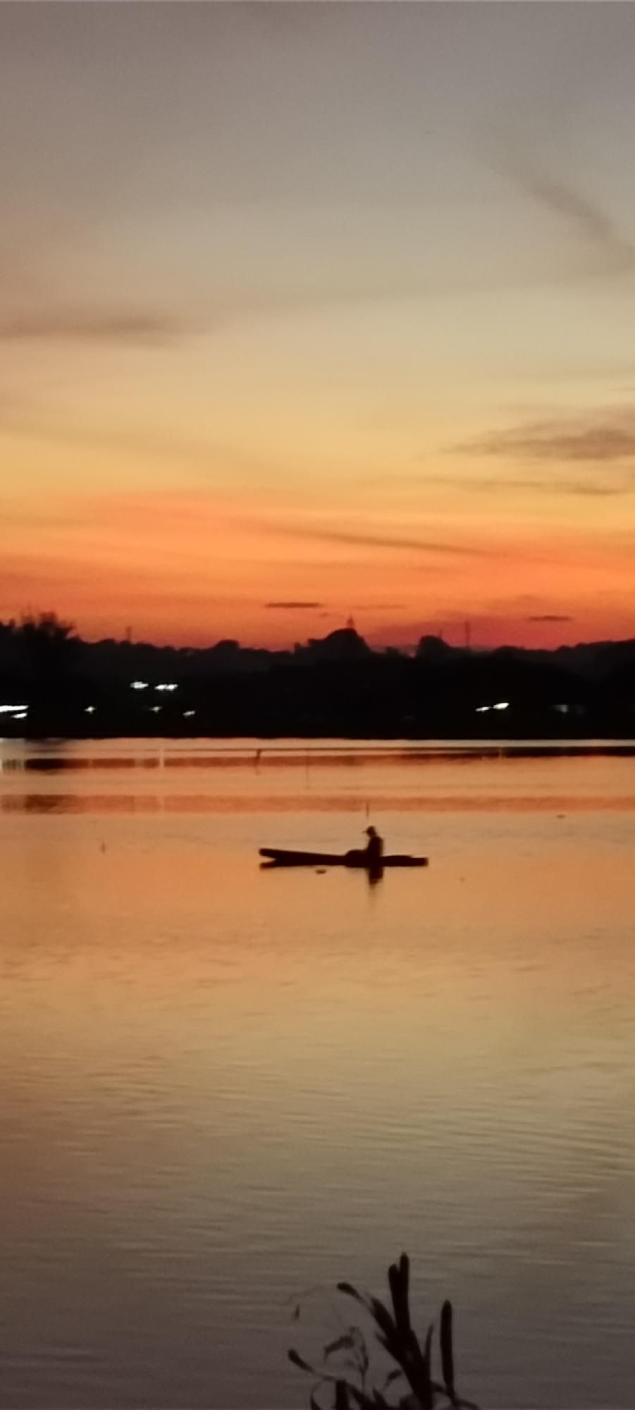 The image depicts a serene sunset scene over a body of water, likely a lake or river, in Lhokseumawe, Indonesia. The sky is a gradient of warm colors, transitioning from a pale gray at the top to vibrant orange and red hues closer to the horizon. Silhouetted against the colorful sky is a distant cityscape, featuring the distinct dome of a mosque and other buildings, some with illuminated windows.
In the foreground, a single individual is seated in a small, dark boat. The boat is long and narrow, appearing to be a canoe or a similar vessel. The person in the boat is also silhouetted, making it impossible to discern any specific features. The water's surface reflects the colors of the sunset, creating a shimmering, orange-toned expanse. Small ripples on the water suggest a gentle breeze. In the lower right corner, the dark silhouette of some aquatic plants or reeds adds a touch of natural detail to the foreground. The overall impression is one of tranquility and natural beauty during twilight.