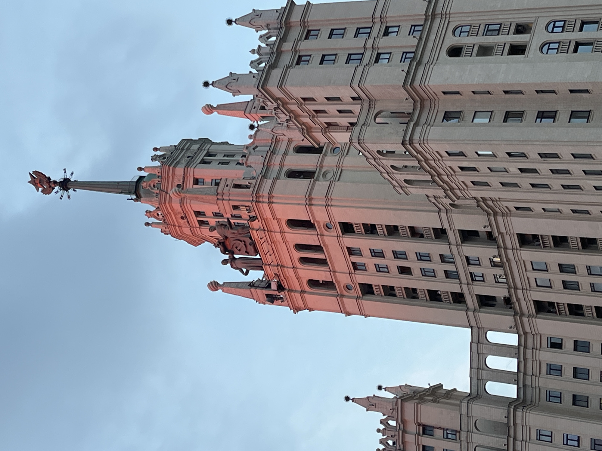 The image shows the Kotelnicheskaya Embankment Building, a Stalinist skyscraper located in Moscow, Russia. The building is illuminated by the warm, rosy glow of sunset. The upper section of the skyscraper, featuring ornate spires and statues, is prominently visible against a pale blue sky with faint clouds. The architectural style is characterized by its grandeur and classical elements. The building's facade is constructed from light-colored stone, and numerous windows are visible, indicating its residential or office use. The image is a close-up view, focusing on the detailed architecture of the upper floors and the spire topped with a star.