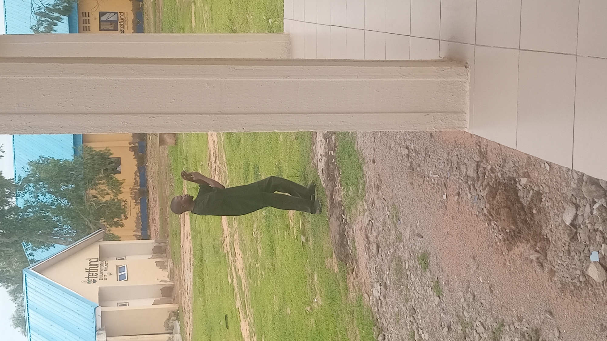 The image shows a person standing outdoors in front of a building. The person is wearing dark clothing and appears to be looking up and to the right. The setting seems to be a rural or semi-urban area, with a grassy field and a dirt path visible. In the background, there is a building with a blue roof and yellow walls, featuring some text that appears to read "Tetfund" and "2017 Project". The weather appears to be clear, and the time of day is likely daytime. The image is oriented vertically, with the person standing on the right side. The overall impression is one of a casual outdoor scene.