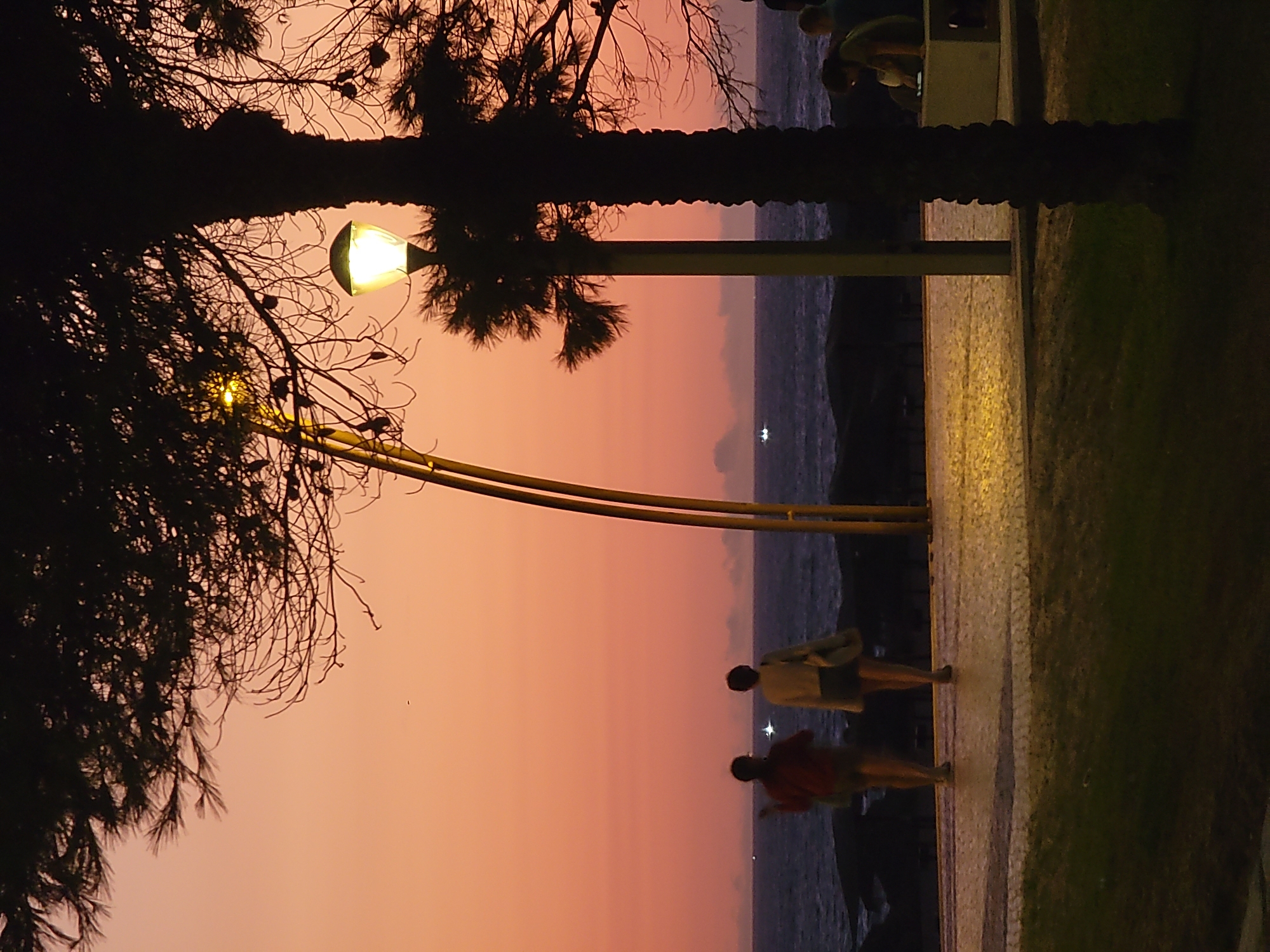 The image depicts a sunset scene at the Quarteira waterfront in Portugal. Two individuals are walking away from the camera on a cobblestone promenade. The sky is a gradient of orange and pink, and the ocean is visible in the background with gentle waves. Palm trees and other vegetation cast shadows across the scene, with a lit lamppost adding illumination to the promenade. Further down the promenade, another person is seated on a bench. The overall atmosphere is tranquil and serene.