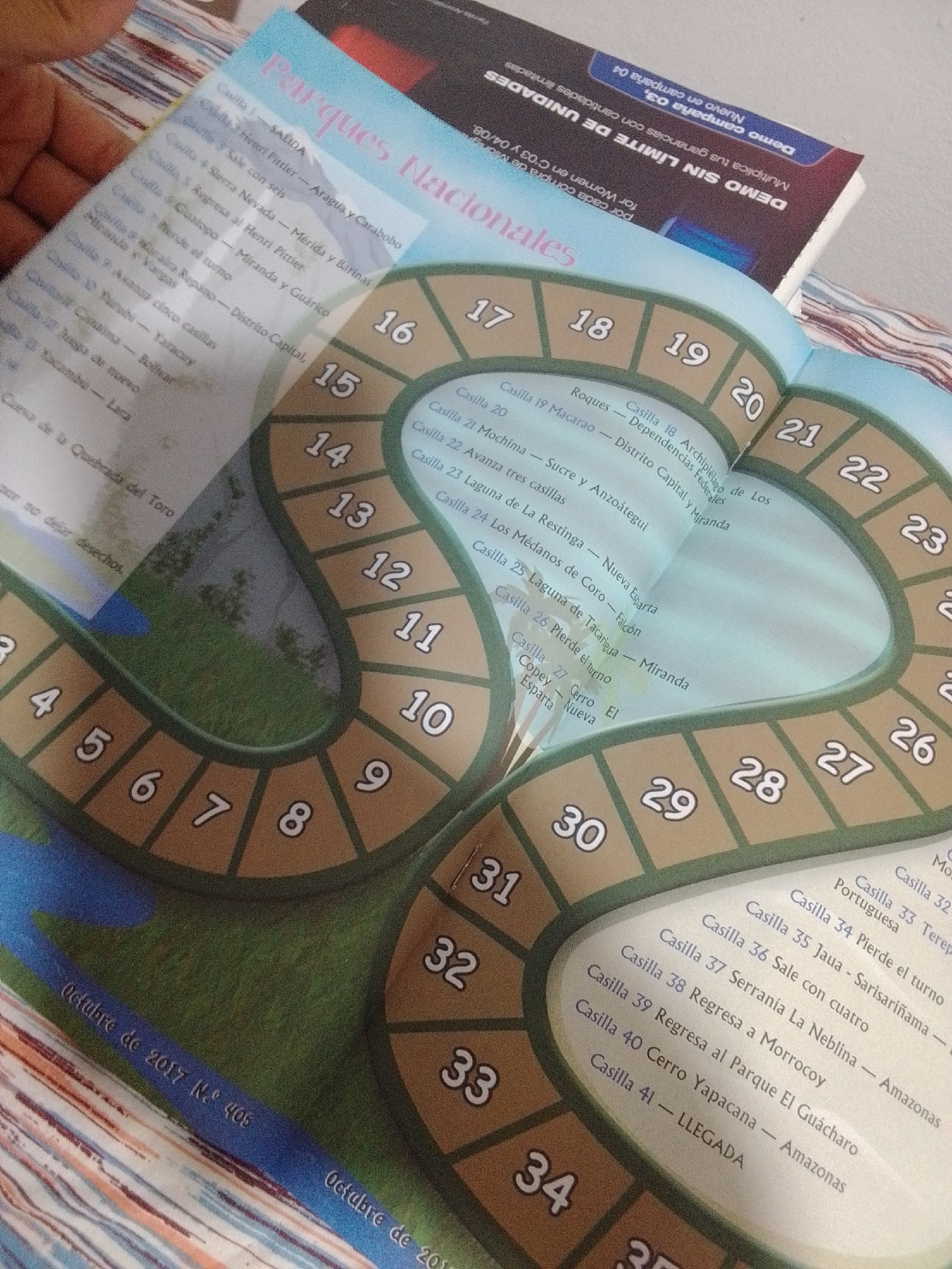 The image shows a close-up of an open board game with a winding path of numbered spaces. The game appears to be themed around Venezuelan National Parks, as indicated by the title "Parques Nacionales" at the top. The spaces are marked with numbers from 1 to at least 41. Text listing park names and locations is visible next to some of the numbered spaces, such as "Casilla 18 Archipiélago de Los Roques - Dependencias Federales" and "Casilla 33 Terepaima - Portuguesa." A blue, winding river graphic is depicted in the lower portion of the game board, and grassy areas with trees are visible within the path. The lower edge of the game board has text that reads "Octubre de 2017 N.º 405". A book with text in Spanish, possibly related to a promotion or advertisement, is partially visible behind the board game. The scene is lit from above and slightly to the left. There are no people clearly visible in the frame. The provided crops offer clearer views of different sections of the board game and the text. The image does not provide any specific information about the city of La Tagua, Venezuela.