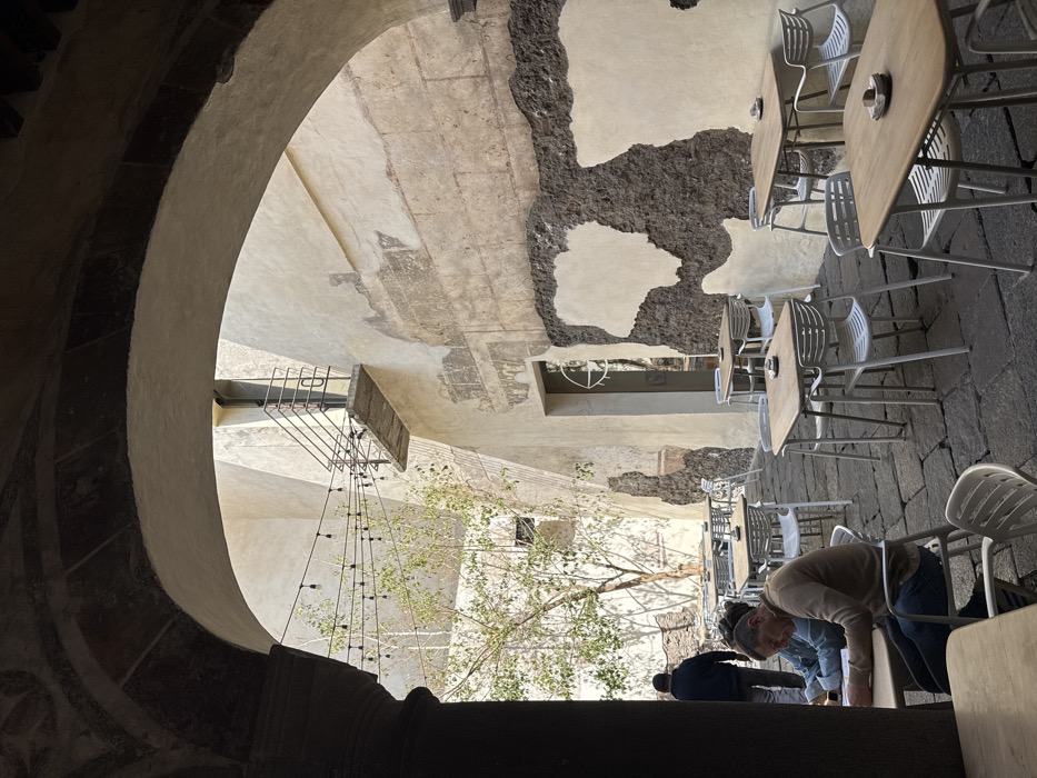The image depicts an open-air courtyard cafe, viewed through a dark, stone arched passageway, located within a historic building in Puebla, Mexico. The courtyard floor is paved with dark, irregular cobblestones. Tall, aged walls enclose the space; they are primarily light cream or off-white, with significant sections of plaster missing, revealing darker, rougher stone or brickwork beneath. Faint rectangular outlines, possibly remnants of older frescoes or architectural details, are visible on some plastered sections.
In the mid-ground, a leafless tree with light-colored bark stands to the left, casting shadows. String lights with bare bulbs are strung horizontally across the upper portion of the courtyard. A small, cantilevered concrete balcony with a dark metal railing extends from an upper-story wall on the right. Below it, a modern glass-paneled door with a circular white logo featuring a crosshairs-like design is set into the wall.
Numerous square or rectangular tables with light wood tops and white, modern-style plastic chairs are arranged throughout the courtyard. Some tables have small, dark, circular objects on them, possibly ashtrays or condiment holders.
Three individuals are visible. In the foreground-left, a man in a brown long-sleeved shirt and blue jeans is seated at a table, leaning forward, engaged in reading or writing in a yellow book. A second person is partially visible next to him, wearing a light blue denim jacket. Further into the background, a third man in a dark blue hooded sweatshirt walks away from the viewer, deeper into the courtyard. The scene is illuminated by natural daylight, indicating daytime.