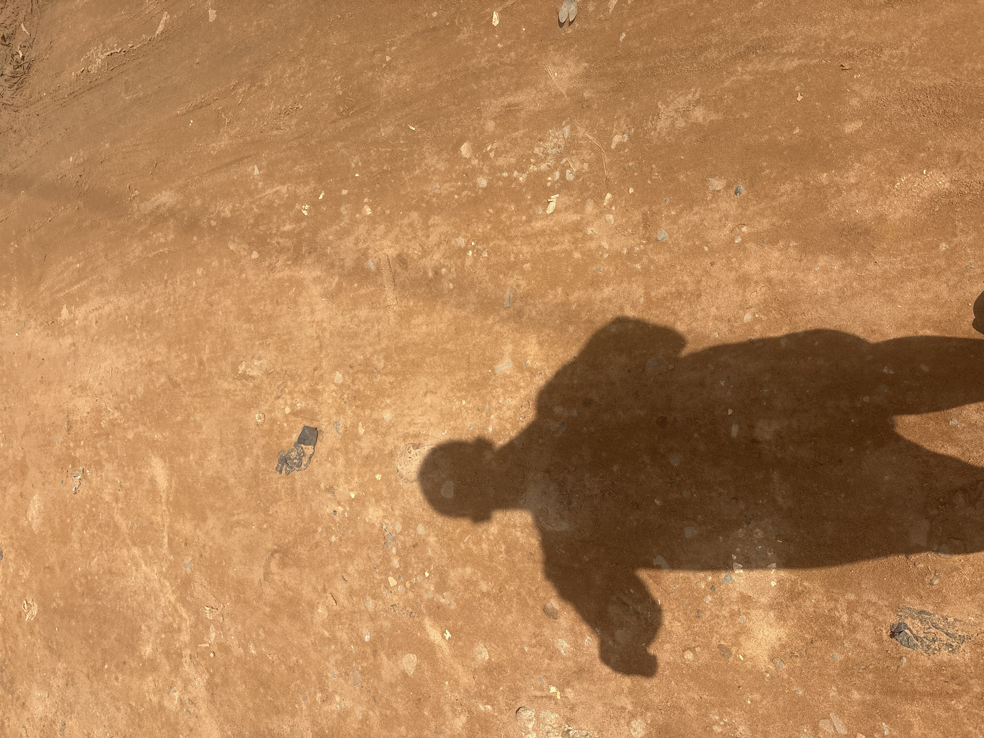 The image shows a shadow of a person standing on a dusty, reddish-brown dirt surface in Nkpor, Nigeria. The ground is uneven and appears to be a path or road, with small stones and debris scattered across it. The shadow is cast by a person who is likely holding a phone or camera to take the photo. The lighting suggests it is daytime, with the sun casting a strong shadow. The environment appears to be outdoors, possibly in a rural or less developed urban area.