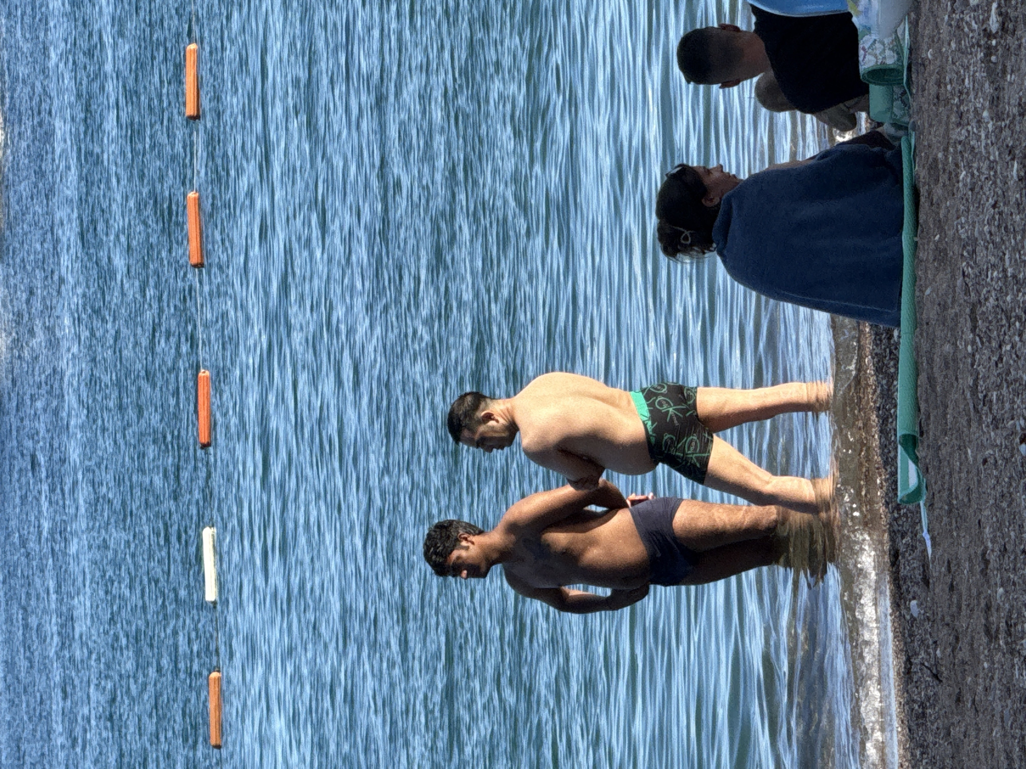 This image depicts a coastal scene in Budva, Montenegro, featuring a body of water and a pebbly beach.
In the upper two-thirds of the frame, calm blue water extends from the top edge to the shoreline. Six rectangular buoys float in a horizontal line across the mid-ground of the water. From left to right, the sequence is orange, white, orange, orange, orange, orange. These buoys appear to be connected by a submerged line, marking a designated area.
Two adult males are standing in the shallow water near the shore, with the water reaching approximately mid-thigh. The male on the left has dark hair and a darker skin tone, wearing dark blue swim briefs. He stands with his hands on his hips, facing right, looking towards the left side of the frame. The male on the right has shorter dark hair and a lighter skin tone, wearing black swim trunks with a green patterned waistband. He stands facing left, looking towards the right side of the frame. Both are positioned with their backs partially to the camera.
The foreground consists of a pebbly beach. Small waves gently break at the water's edge. On the right side of the beach, three individuals are seated. One adult female, with dark hair tied up, is seen from behind, wrapped in a dark blue towel or garment, looking towards the right. To her right, an adult male, also seen from behind, with short dark hair, is wearing a dark t-shirt and sitting on a green and white patterned mat. Between the two adults, a small child (infant/toddler) is partially visible, also seated on the patterned mat. A light green mat is laid flat on the pebbles in the immediate foreground, and part of a blue plastic bottle is visible near the seated individuals.
