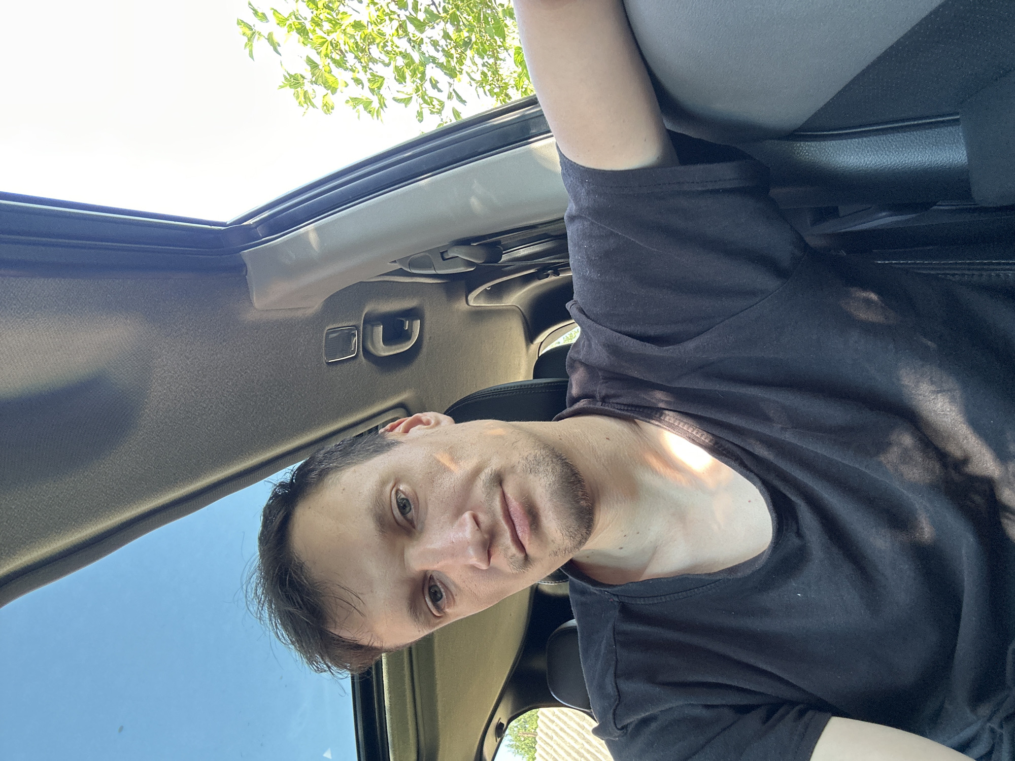 A man with short, dark brown hair and a light beard is taking a selfie in the driver's seat of a car. He is wearing a black t-shirt. Sunlight is streaming in through the sunroof and the open window, casting shadows on his face and chest. The interior of the car is visible, including the dashboard and part of the passenger seat. Outside the car, green trees and a fence are visible. The scene appears to be a sunny day. The location is identified as Shatava, Ukraine, based on the provided information.