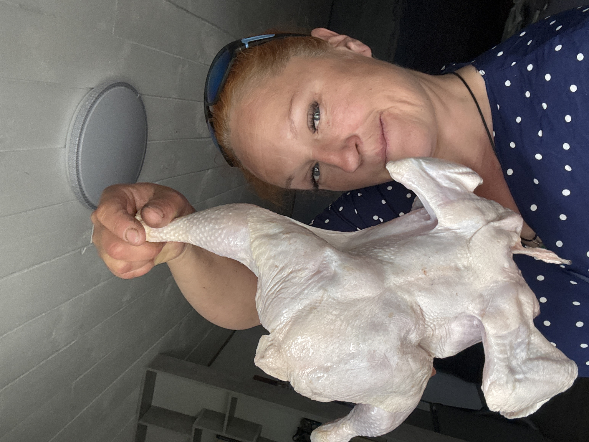 A middle-aged, fair-skinned woman with red hair and blue eyes poses with a raw whole chicken in a room in Chaikovsky, Russia. She wears blue-framed sunglasses atop her head and a navy blue, white-polka-dotted blouse. The woman holds the chicken with her right hand. The background reveals a grey paneled wall with a round, white object attached to it and a white shelving unit.