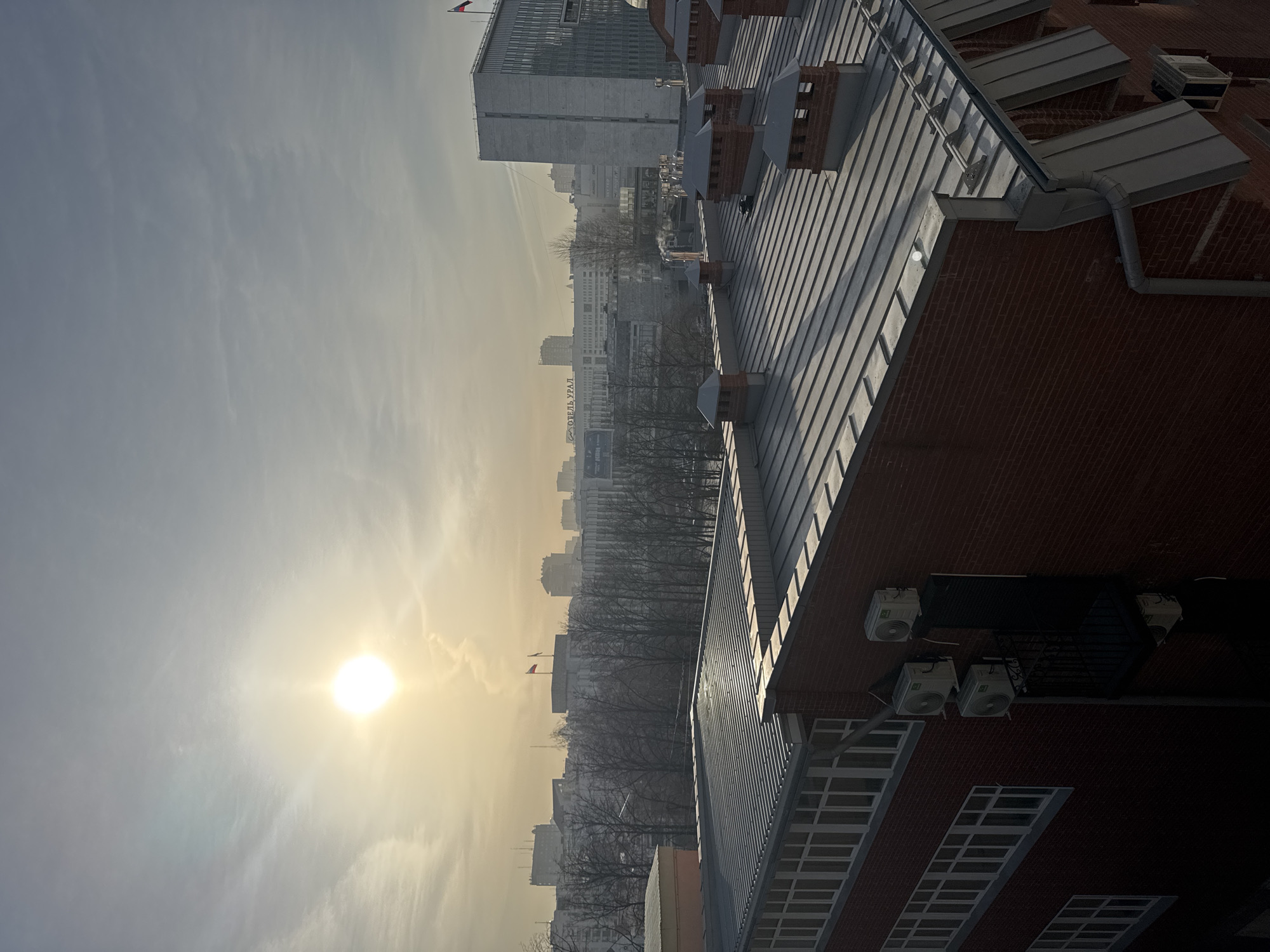 The image shows a cityscape taken from a high vantage point in Perm, Russia. The sun is low in the sky, shining through a hazy atmosphere, creating a warm glow. The cityscape includes several buildings of varying heights and architectural styles, with a line of bare trees in the middle ground. A large brick building with a metal roof dominates the foreground, with multiple air conditioning units attached to its side. The Russian flag can be seen on top of one of the buildings in the distance. A building with the word "ОТЕЛЬ УРАЛ" (Hotel Ural) is visible among the city buildings. The overall impression is one of a cold, sunny day in an urban environment.